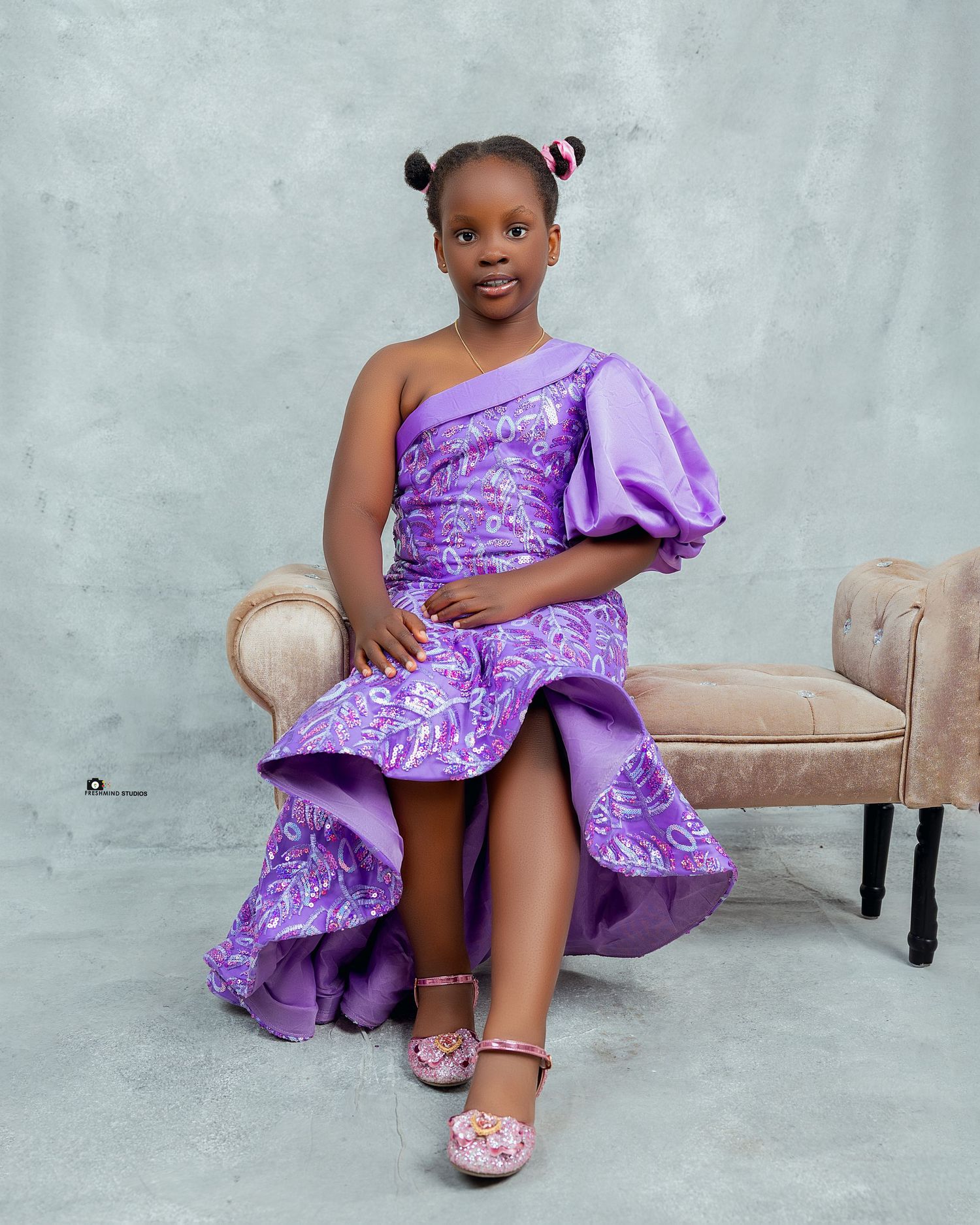 Young model in purple ruffled dress posing elegantly on beige furniture.