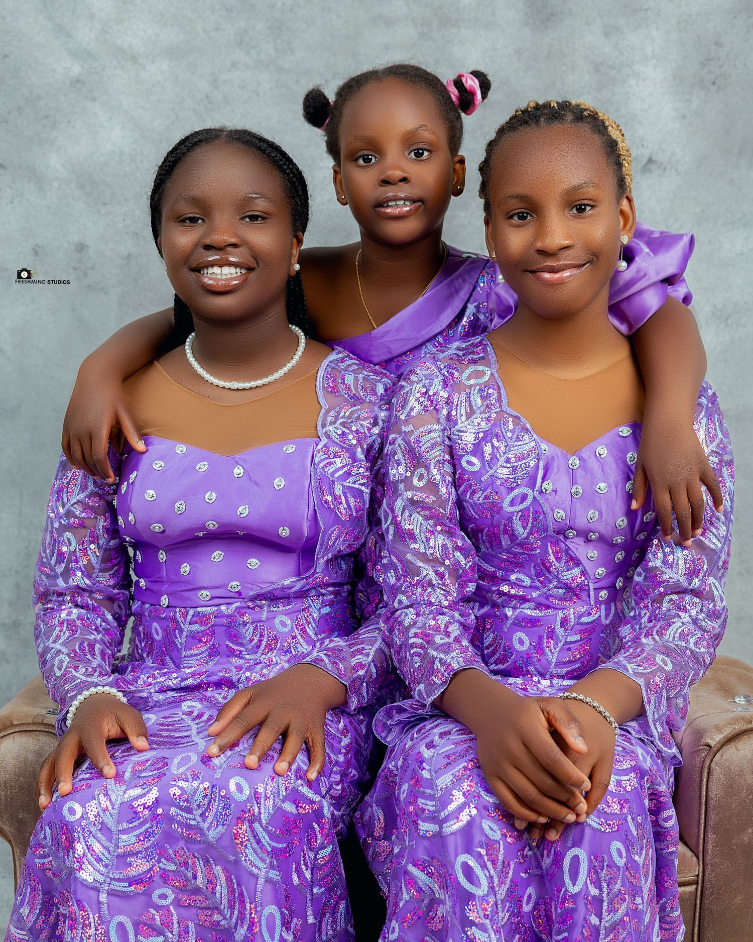 Three young people wearing matching purple traditional dresses with floral patterns posing together.