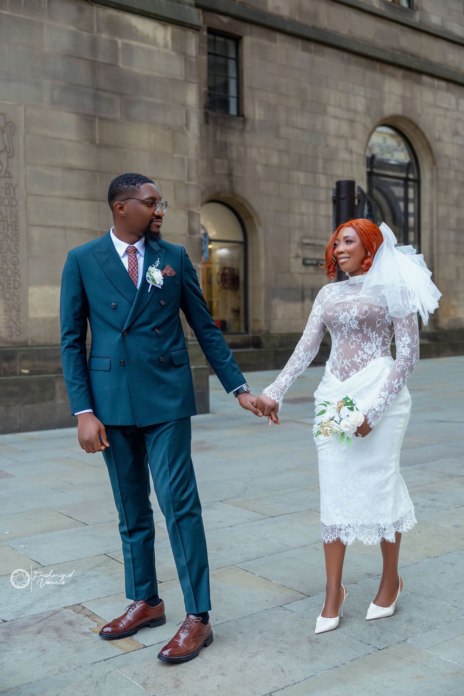 A wedding couple in white lace dress and navy suit pose playfully on stone steps outside a historic building.
