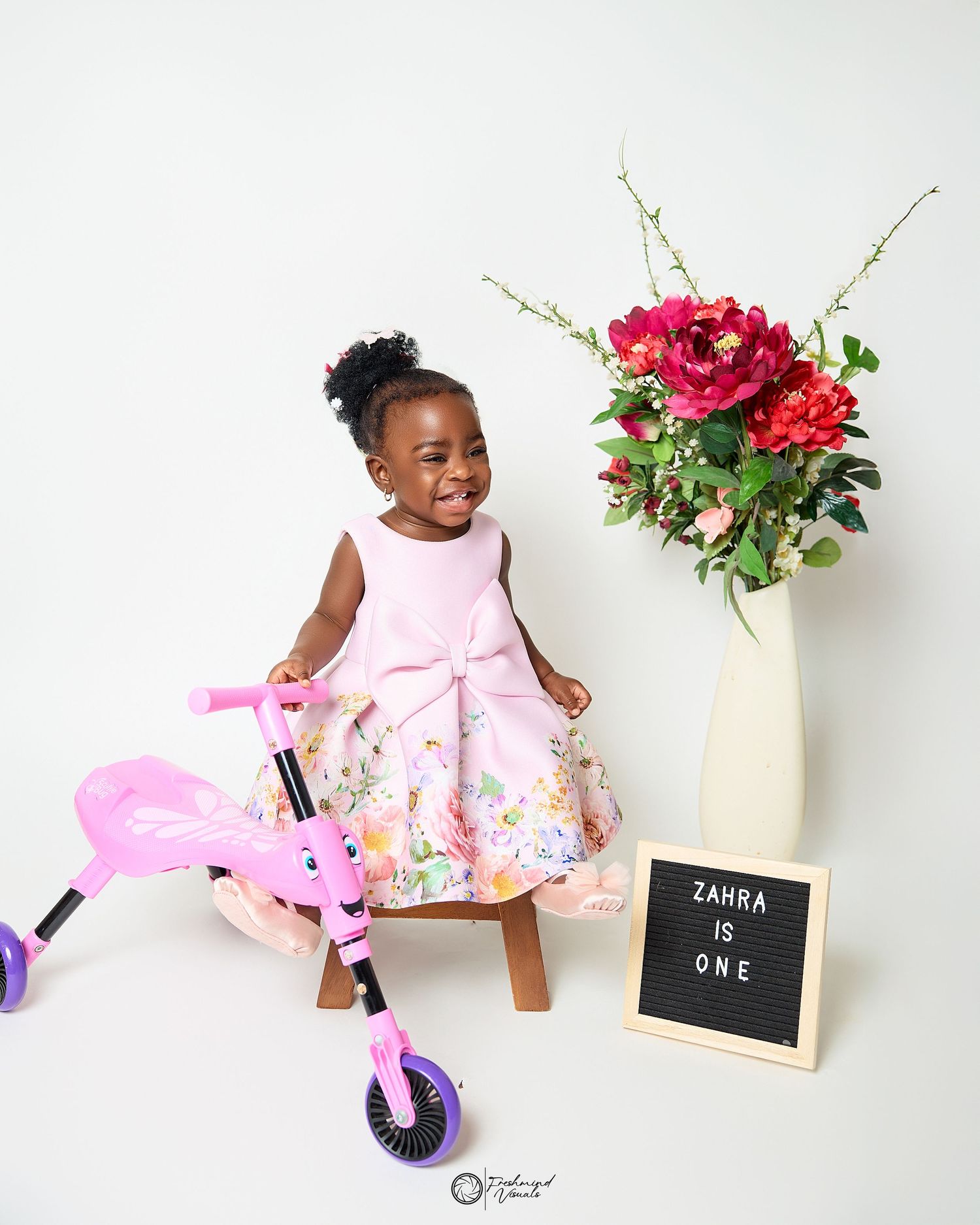 Child on pink balance bike next to floral arrangement and letter board in studio setting.