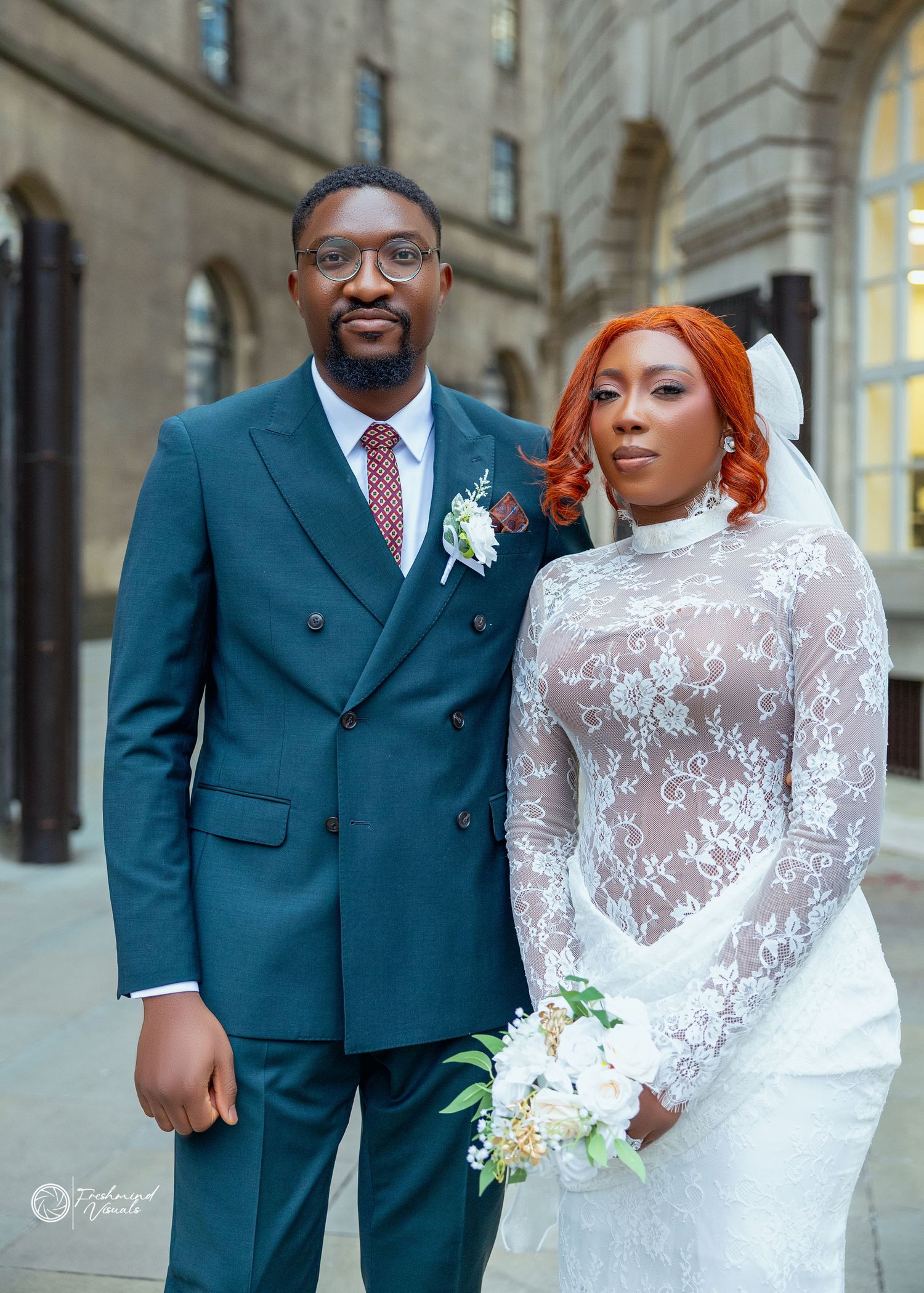 A newlywed couple poses together outdoors, with the bride in a white lace gown and groom in a teal suit.