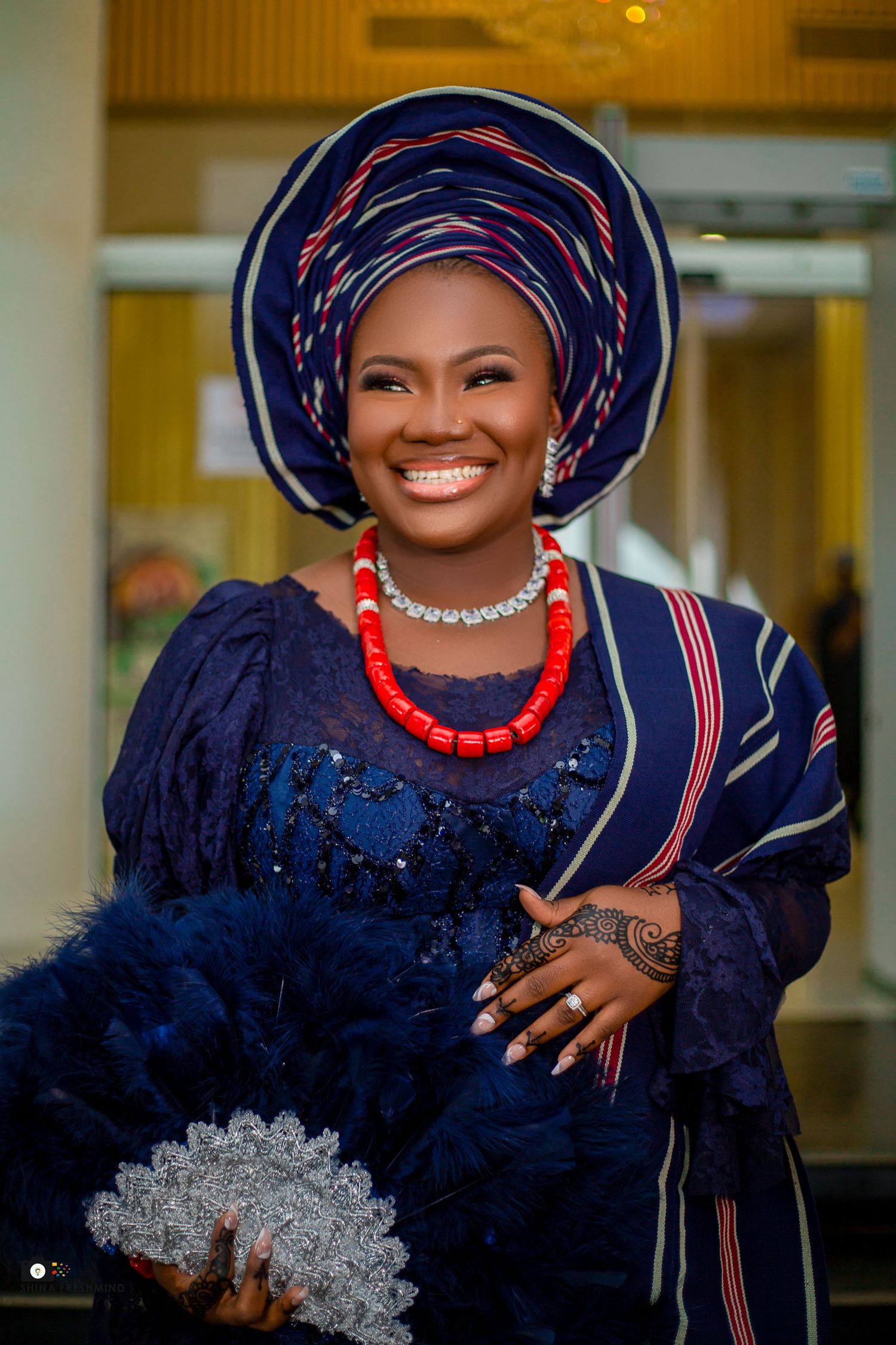 Traditional Nigerian wedding attire featuring navy blue lace dress, gele headwrap, and coral beaded necklace.