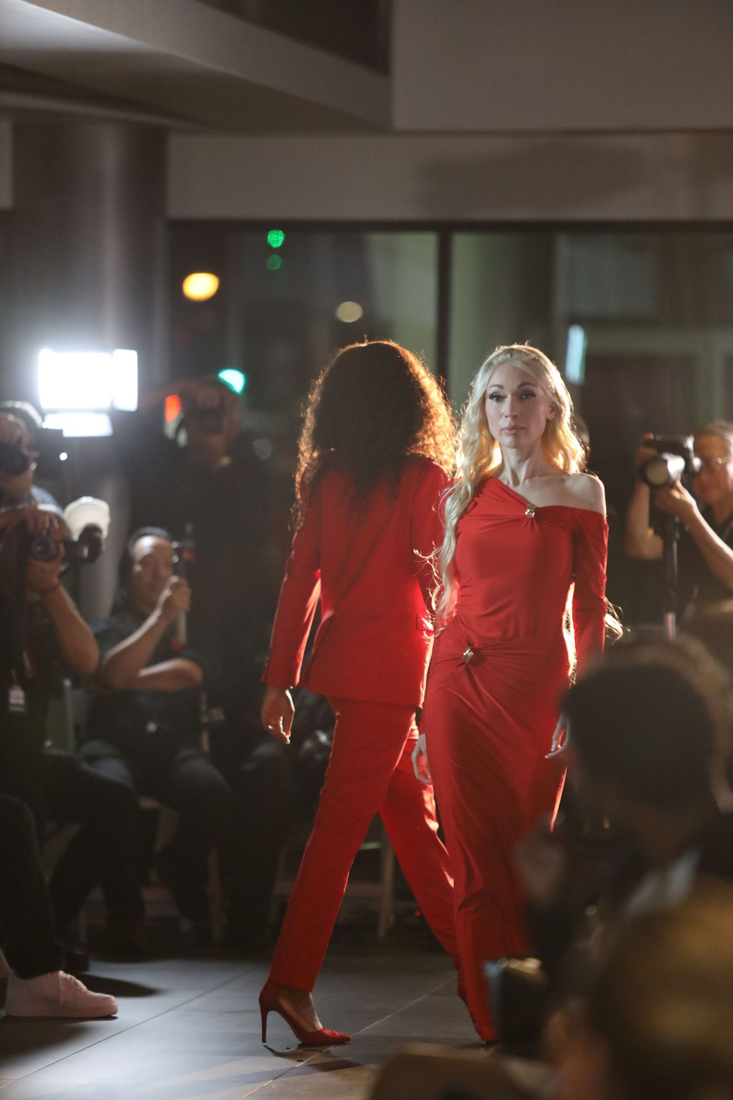 Models in red outfits perform on a dimly lit fashion runway.