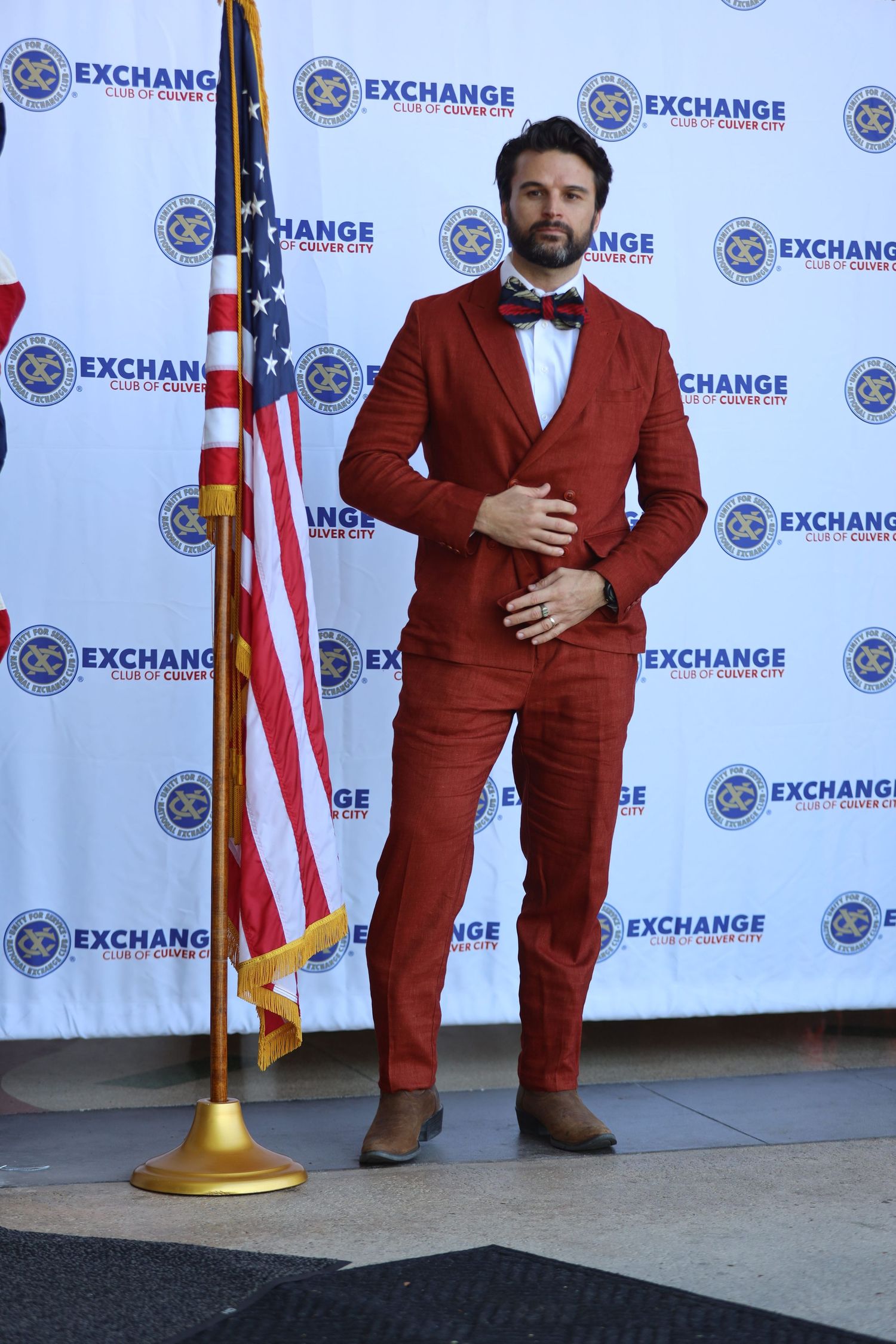 Man in burgundy suit and bowtie poses formally at Exchange event with American flag.