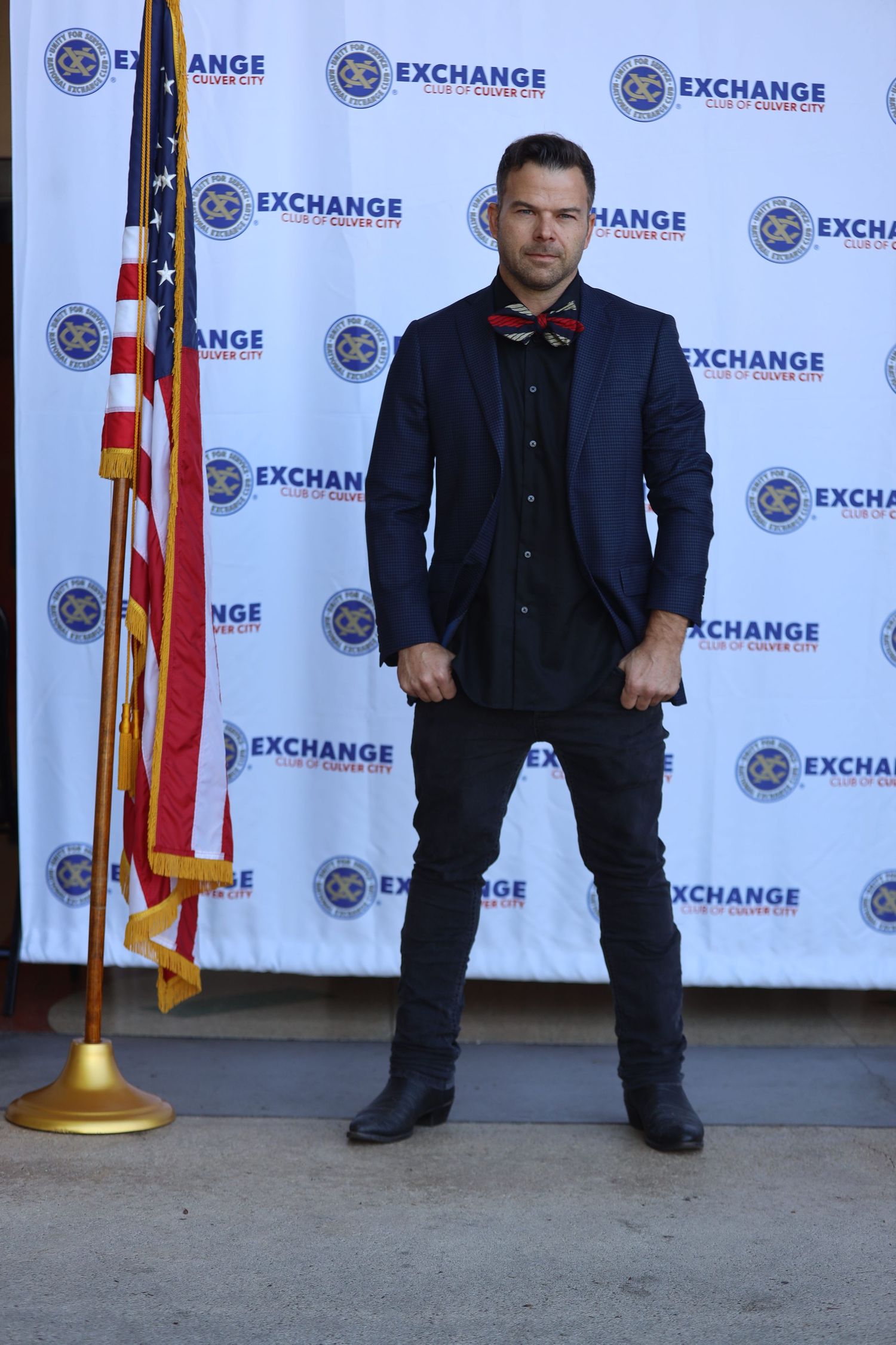 Figure in navy blazer poses against Exchange step and repeat wall.