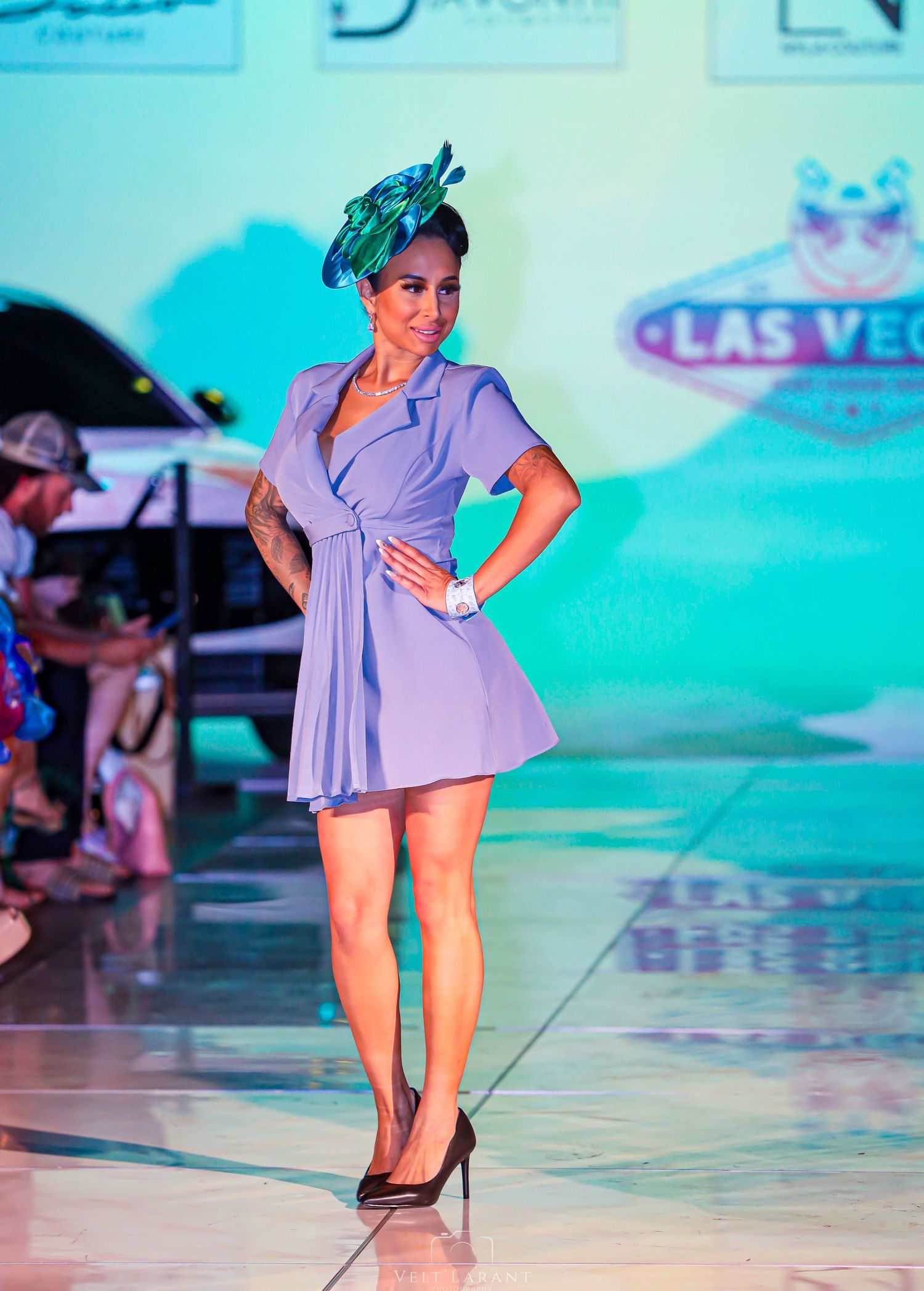 Model in lavender wrap dress and fascinator strikes a pose on runway with Las Vegas backdrop.