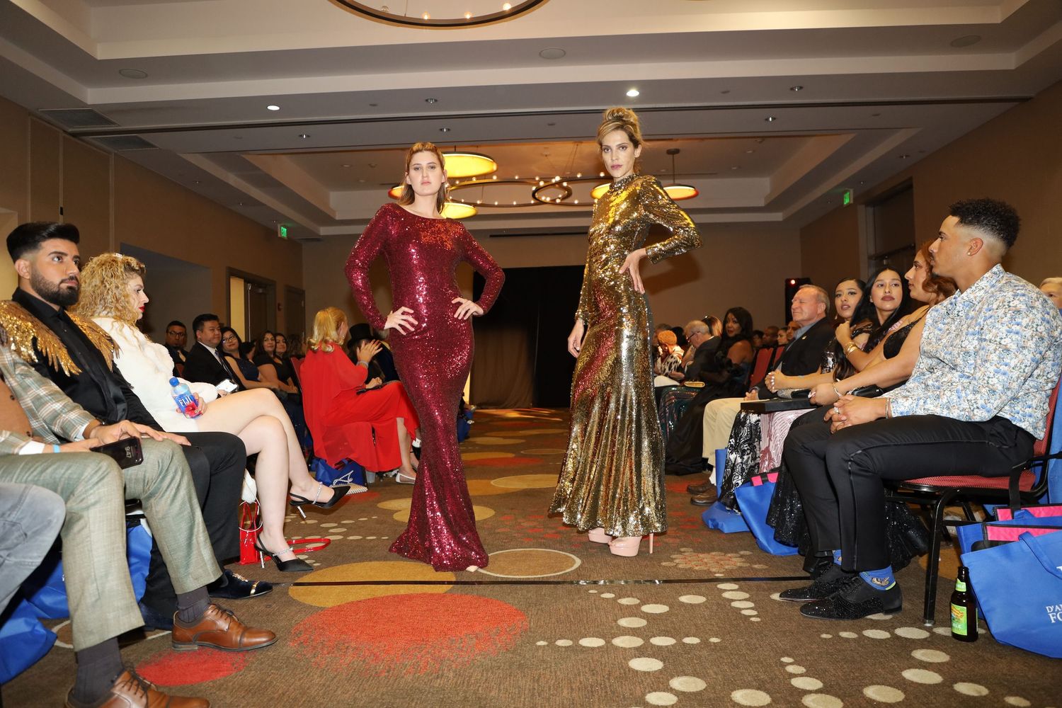 Two models showcase evening gowns on runway as audience watches from both sides.