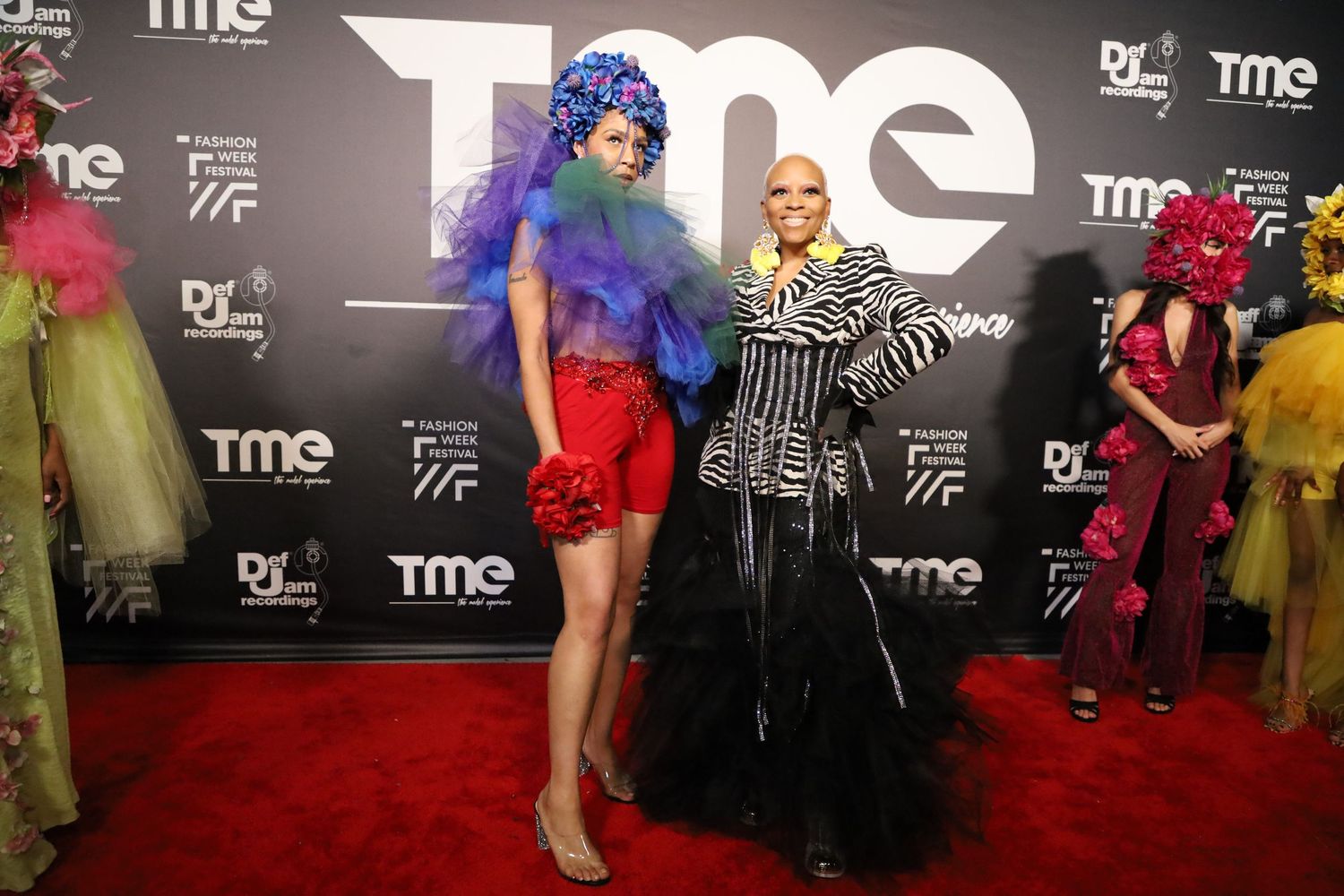 Fashion show attendees in dramatic outfits pose against black TME step-and-repeat wall.
