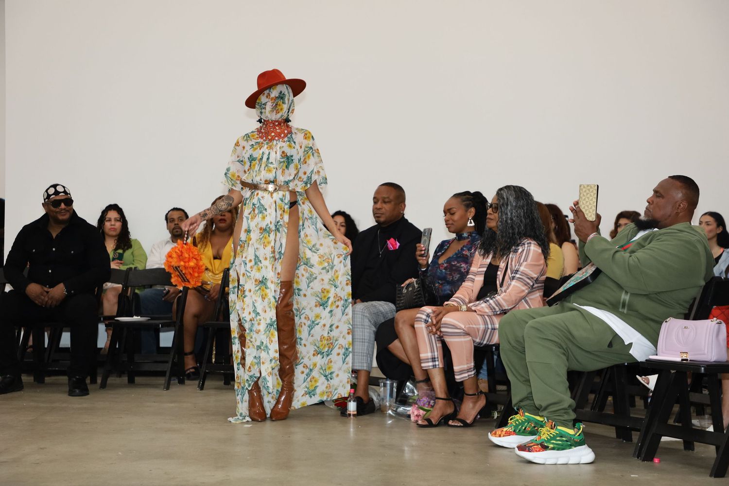 Fashion model walks runway in floral dress and brown boots with red hat at gallery show.