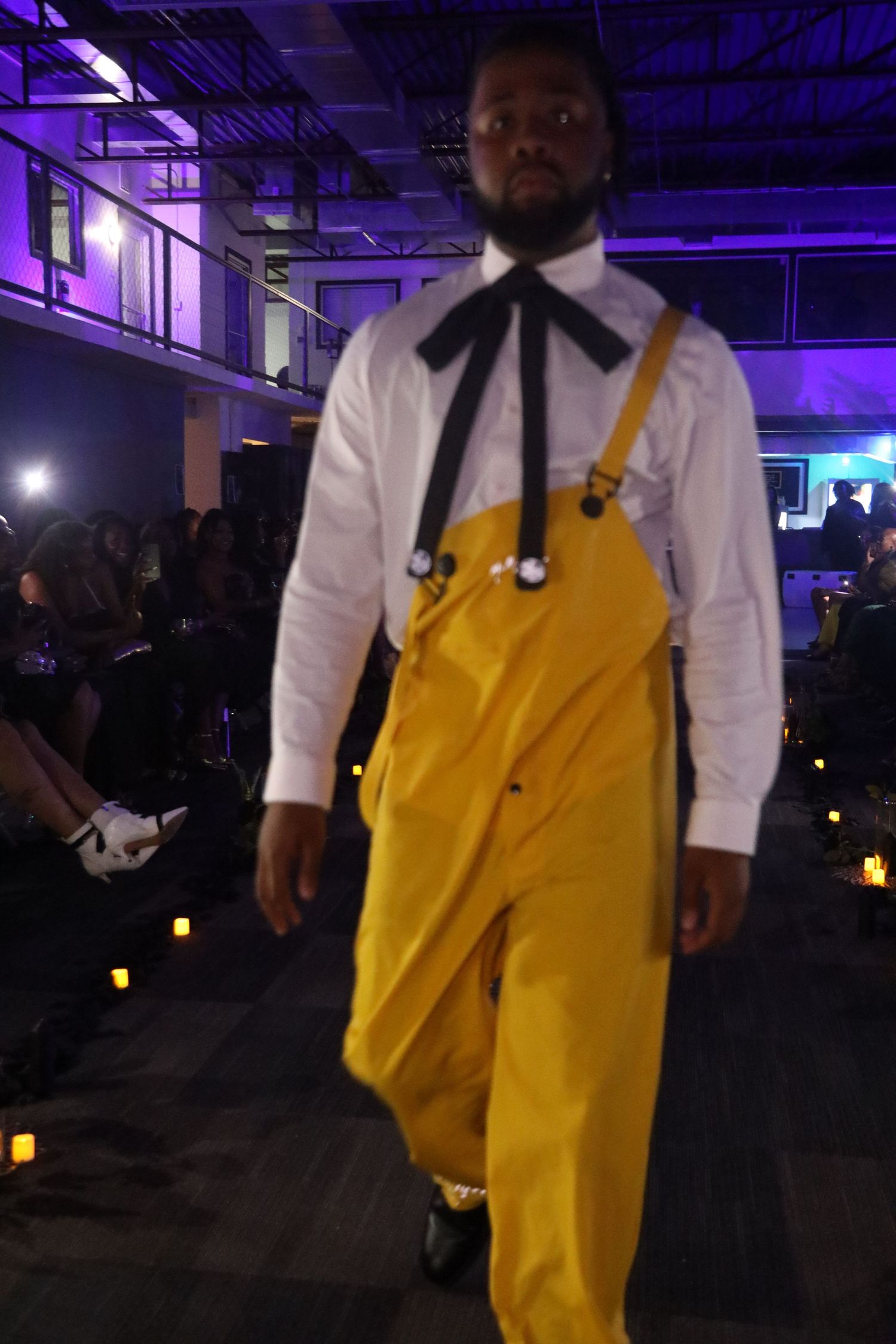 Person in yellow overalls and white shirt standing in dark room with purple ambient lighting.