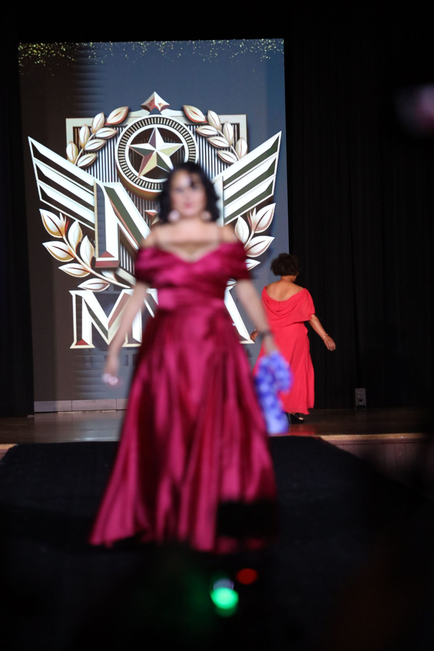 Models in formal gowns walk runway with illuminated logo backdrop.