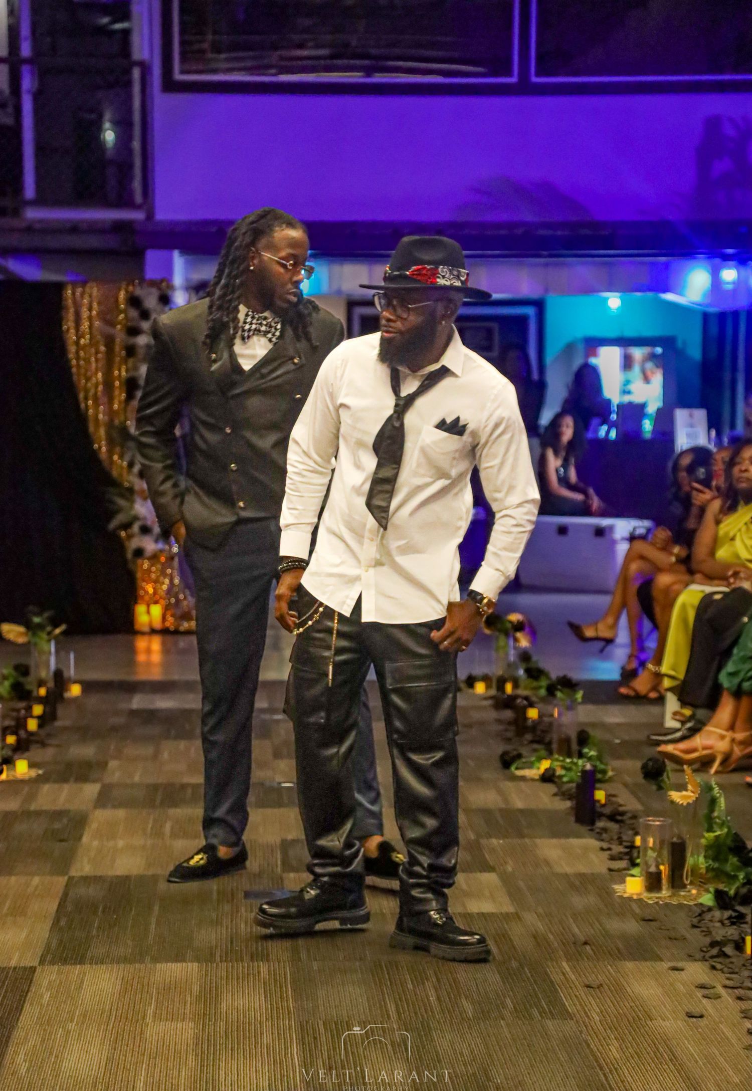 Two models showcase formal menswear on illuminated runway with purple lighting.