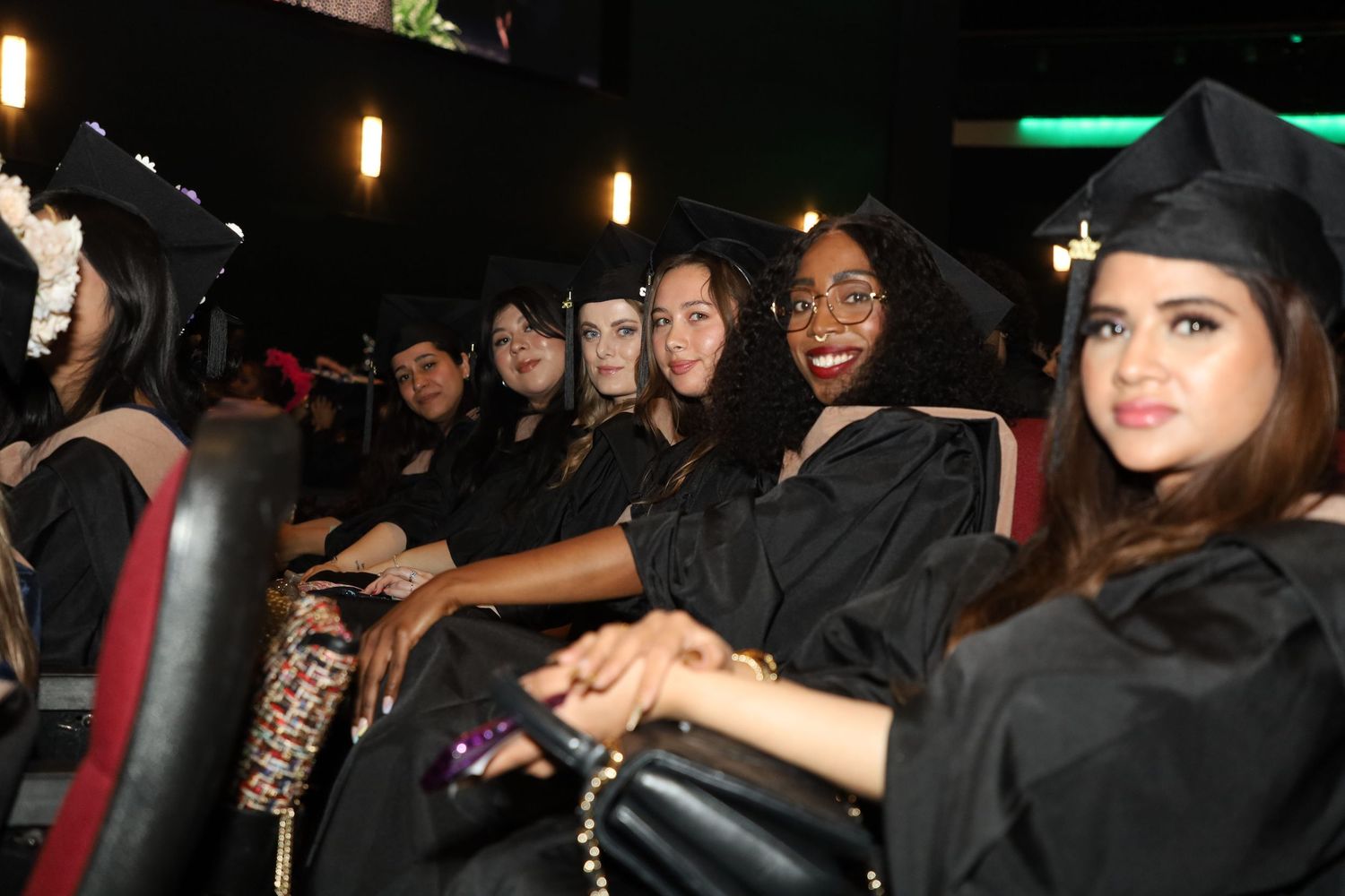 Students in graduation attire seated in theater-style seating with pendant lights overhead.