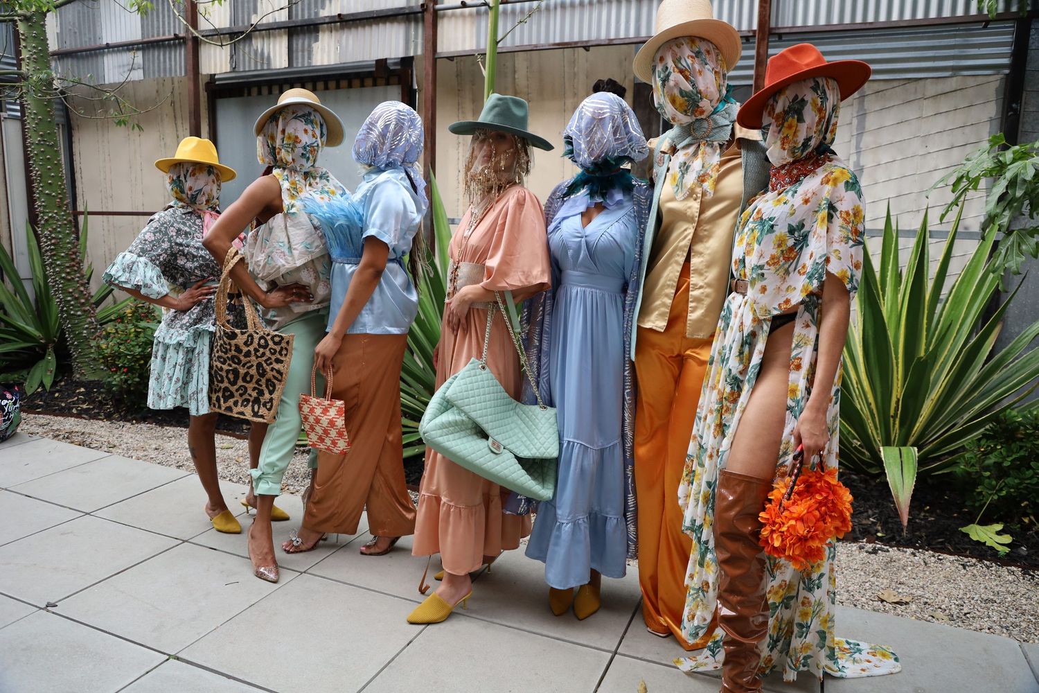 A group poses outdoors wearing colorful vintage-inspired dresses and hats in pastel and earth tones.