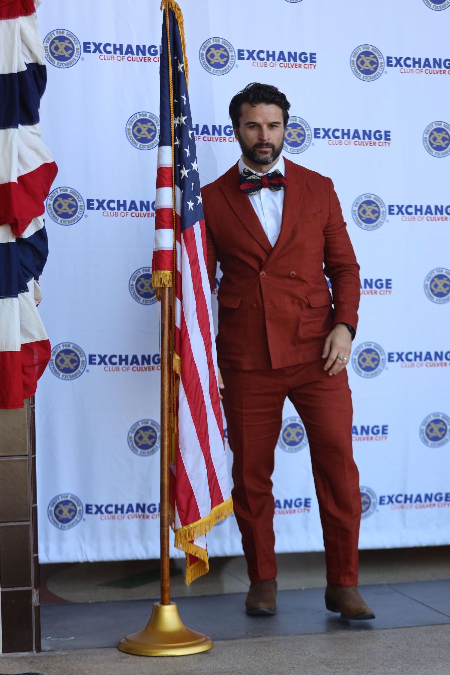 Person in rust-colored suit stands near American flag at corporate event backdrop.