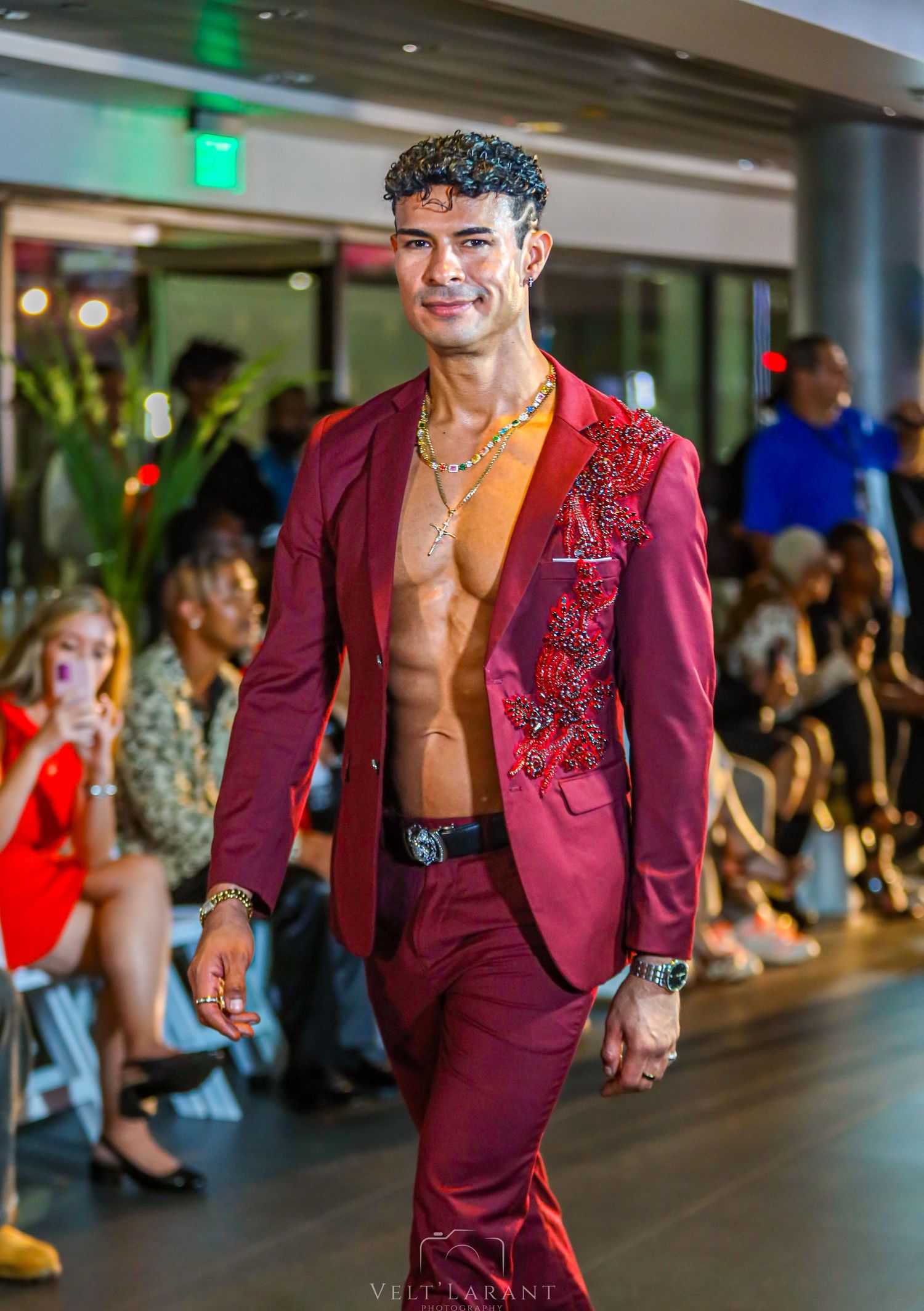 Model presents burgundy suit jacket with embellished details on illuminated runway.