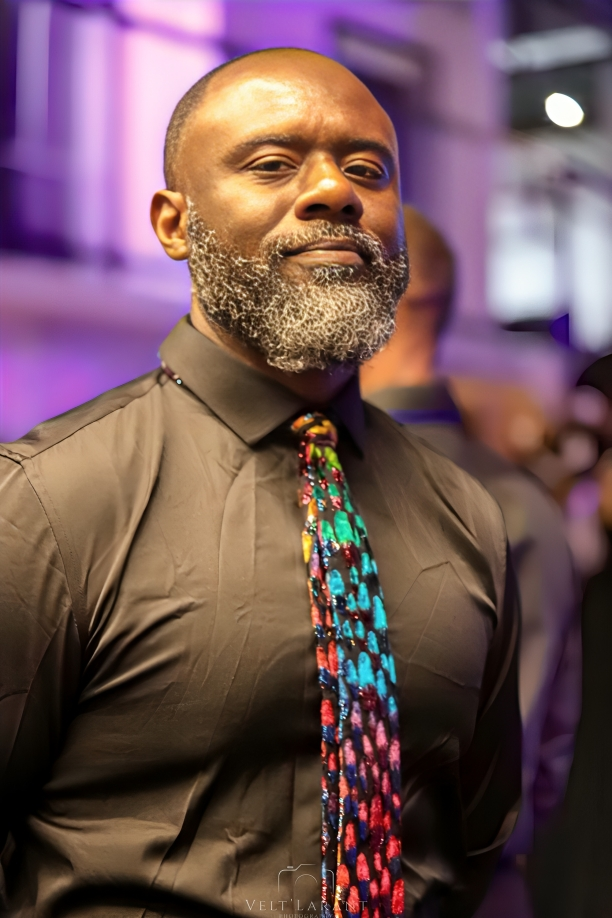 Stylishly dressed man in brown shirt wearing colorful patterned tie in purple-lit environment.