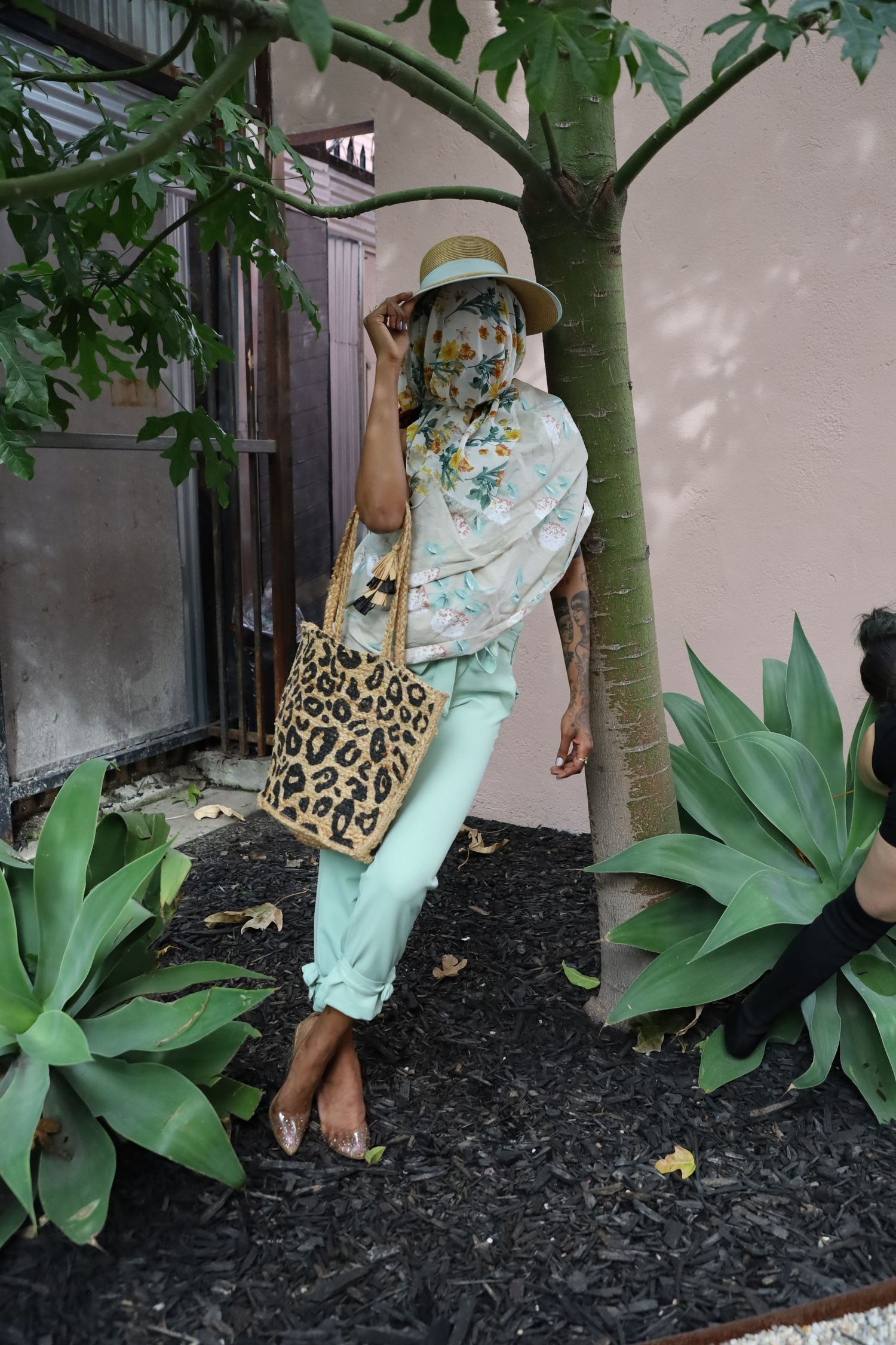 A stylish outfit featuring mint green pants, floral top, straw hat and leopard print bag against tropical foliage.