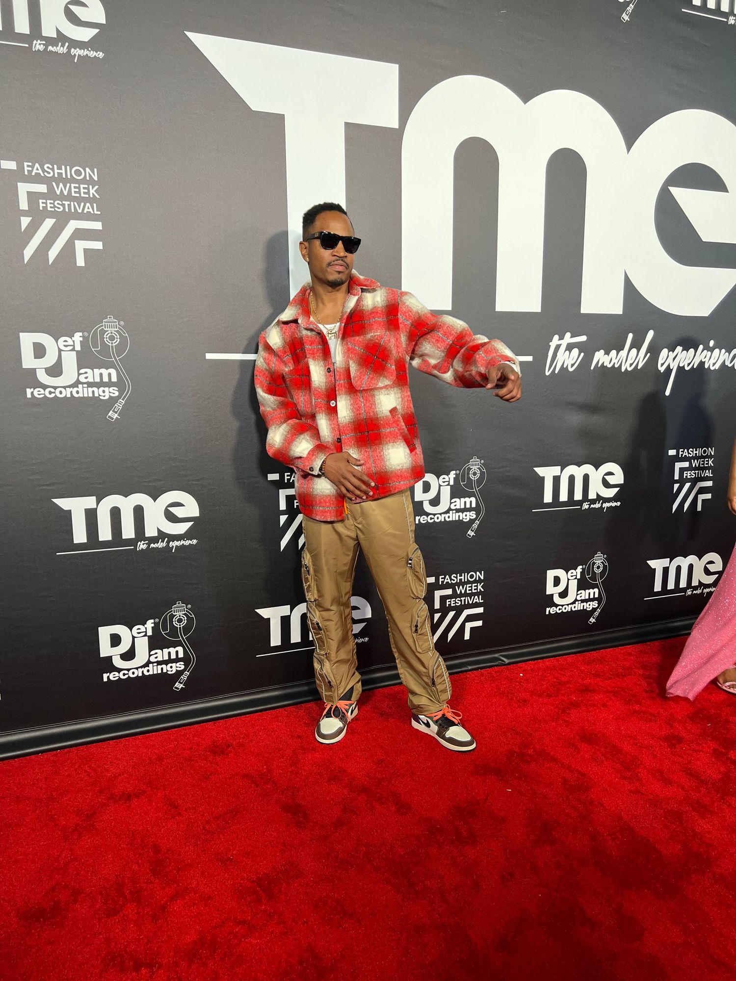 Person in red plaid shirt and khakis poses on red carpet at TME event.