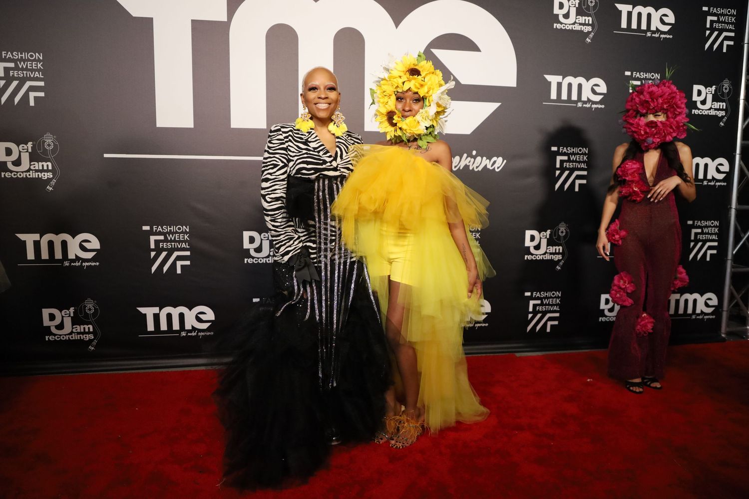Dramatic fashion poses on red carpet with TME branding wall in background.