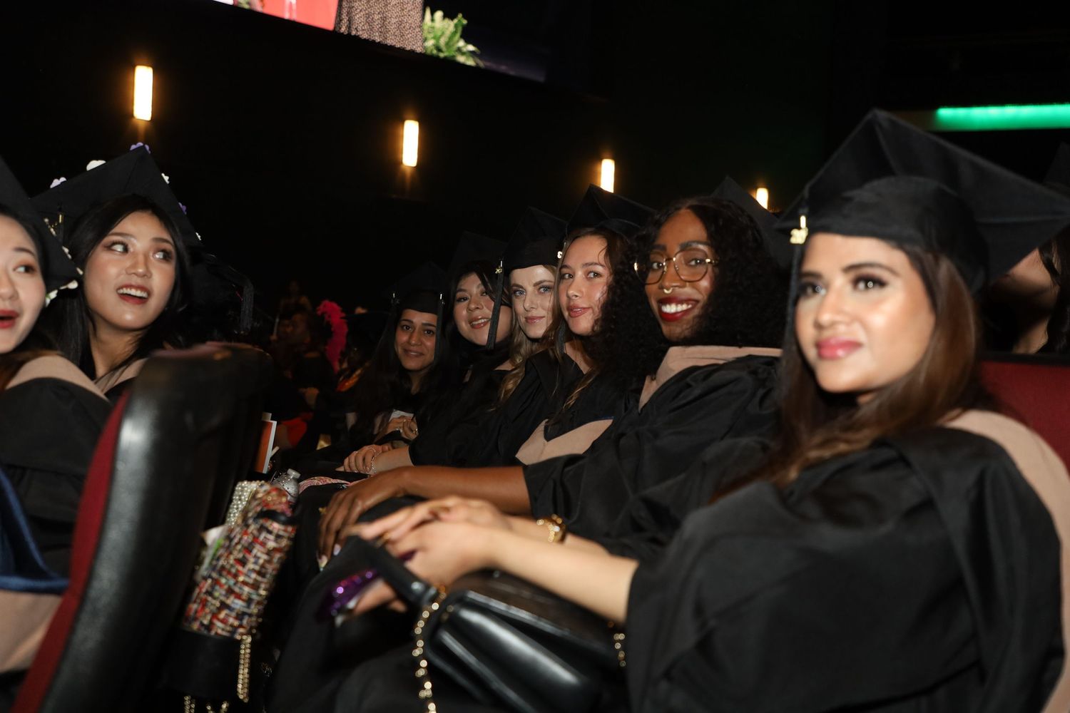 Graduates in black caps and gowns sit together in a dimly lit auditorium during commencement ceremony.