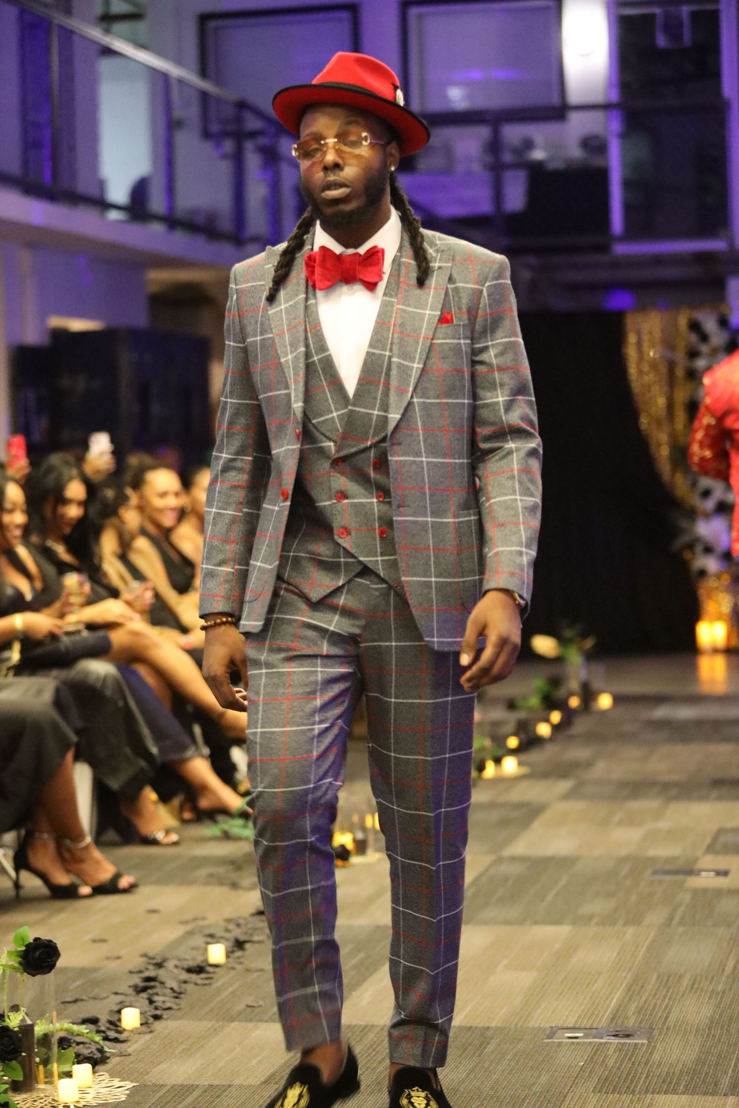 Model wearing plaid suit, red hat and bow tie walks fashion runway.
