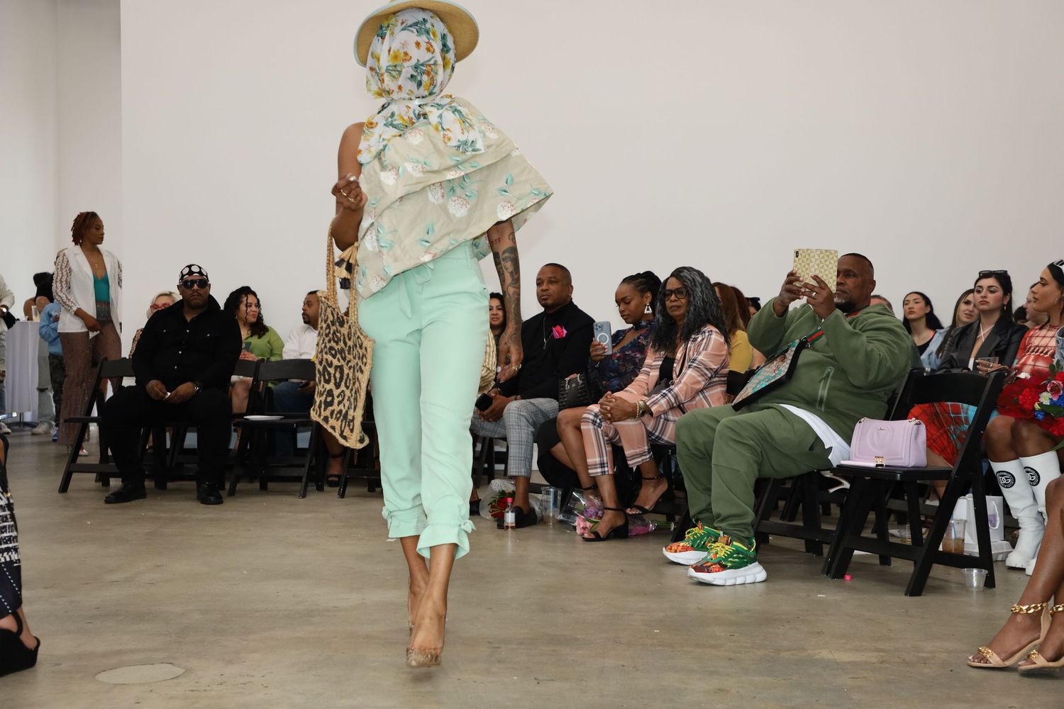 Fashion model wearing mint green pants and floral top strikes dynamic poses on runway with audience in background.
