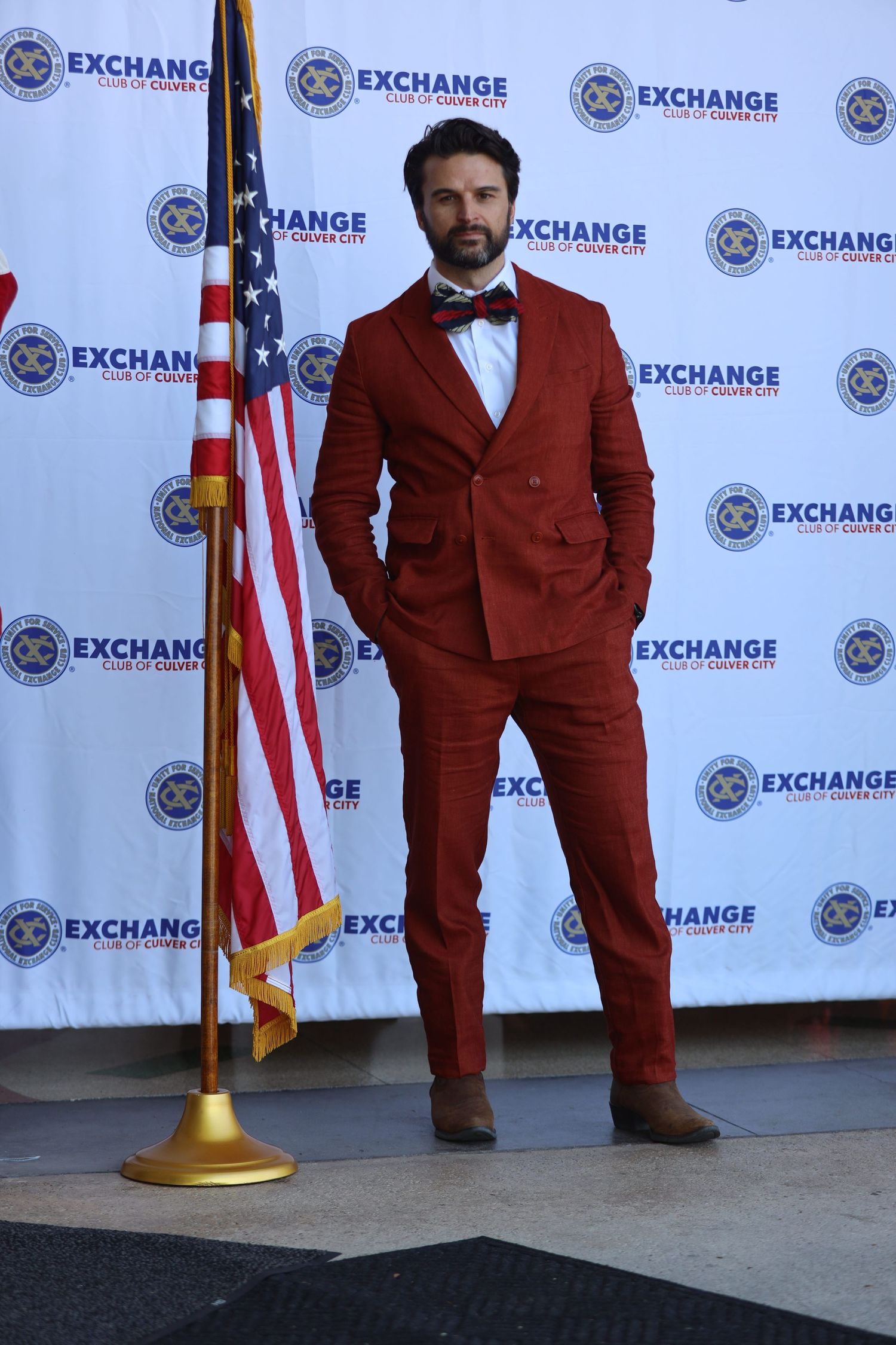 Man in burgundy suit poses confidently next to American flag at Exchange event.