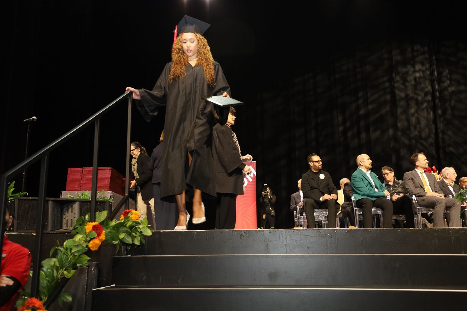 Graduates walk across a dark stage during a formal graduation ceremony.