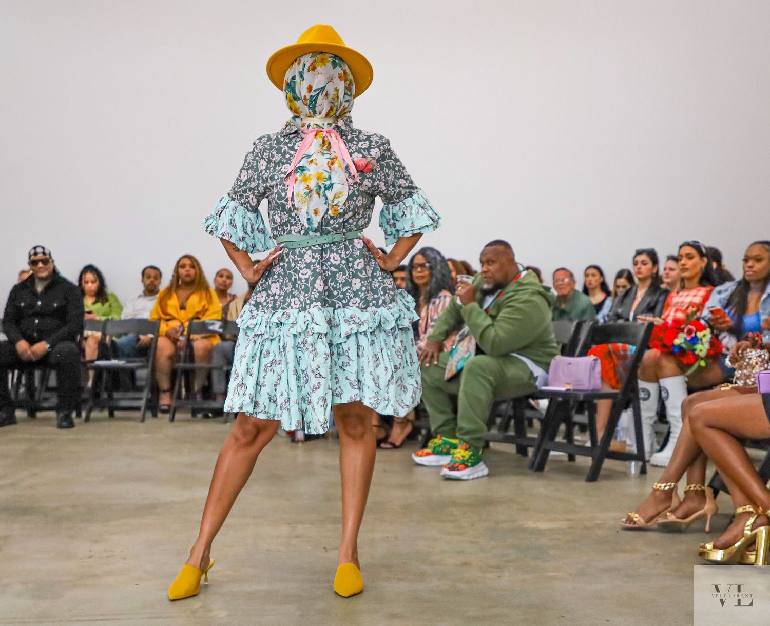 Fashion model showcases yellow hat and floral dress combination on gallery catwalk.