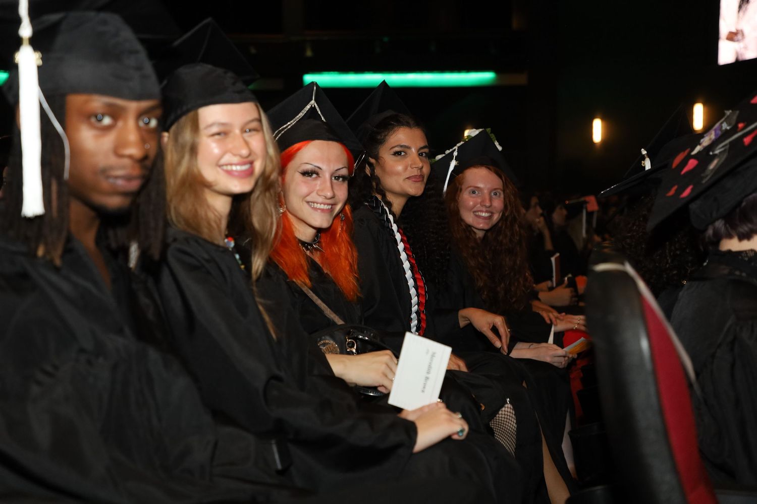 Graduates in black caps and gowns seated together during indoor commencement ceremony.