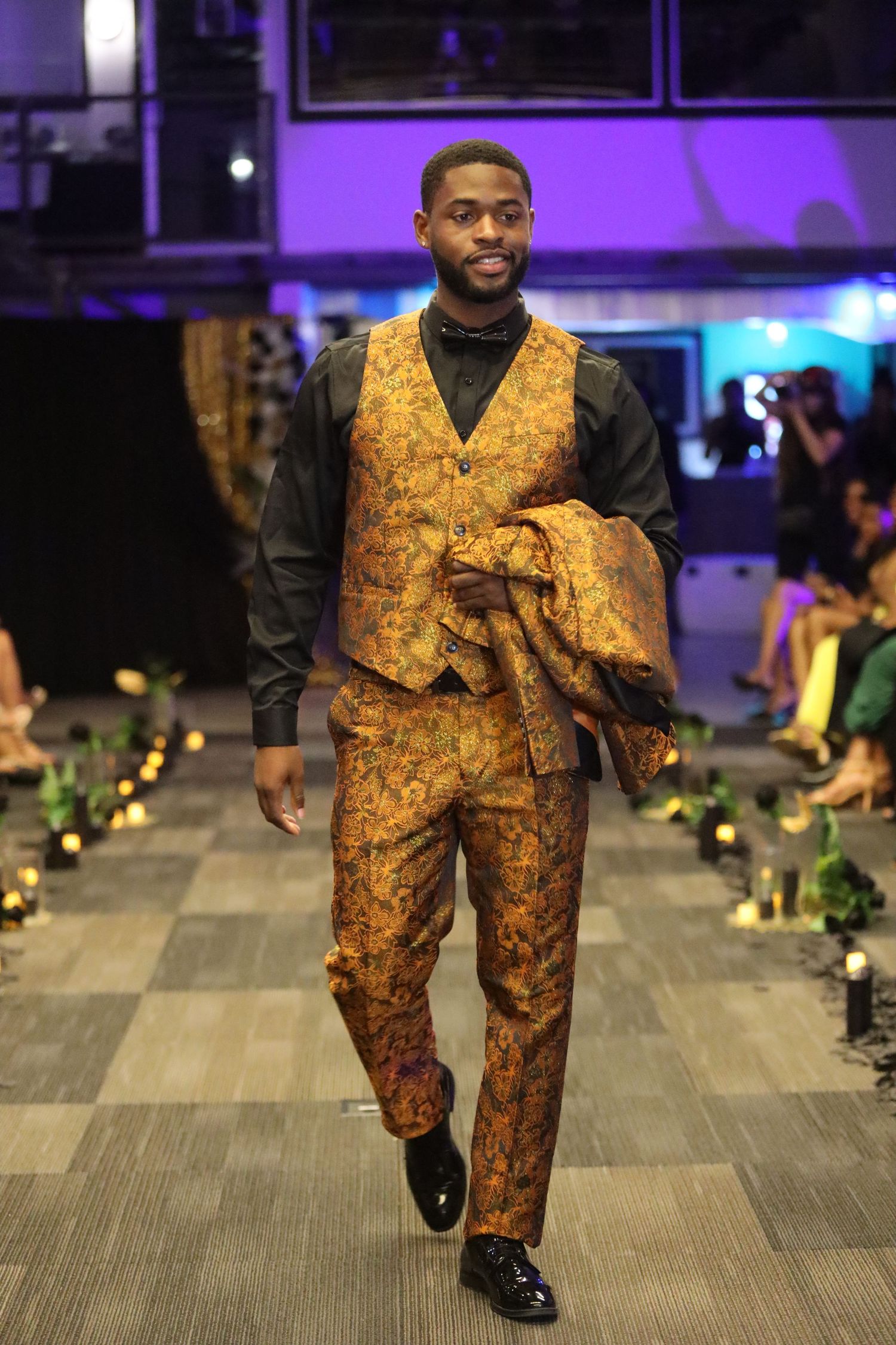 Model walks runway in luxurious gold brocade suit with black shirt at fashion event.