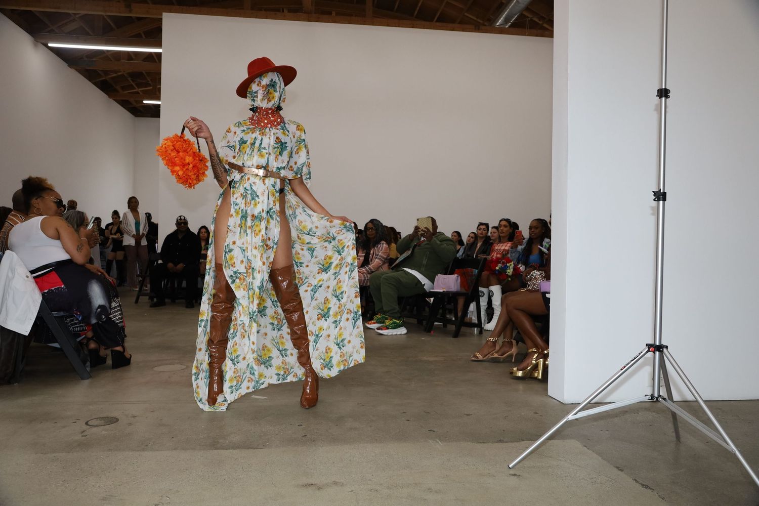 Runway show features flowing floral dress paired with tall brown boots and red hat.