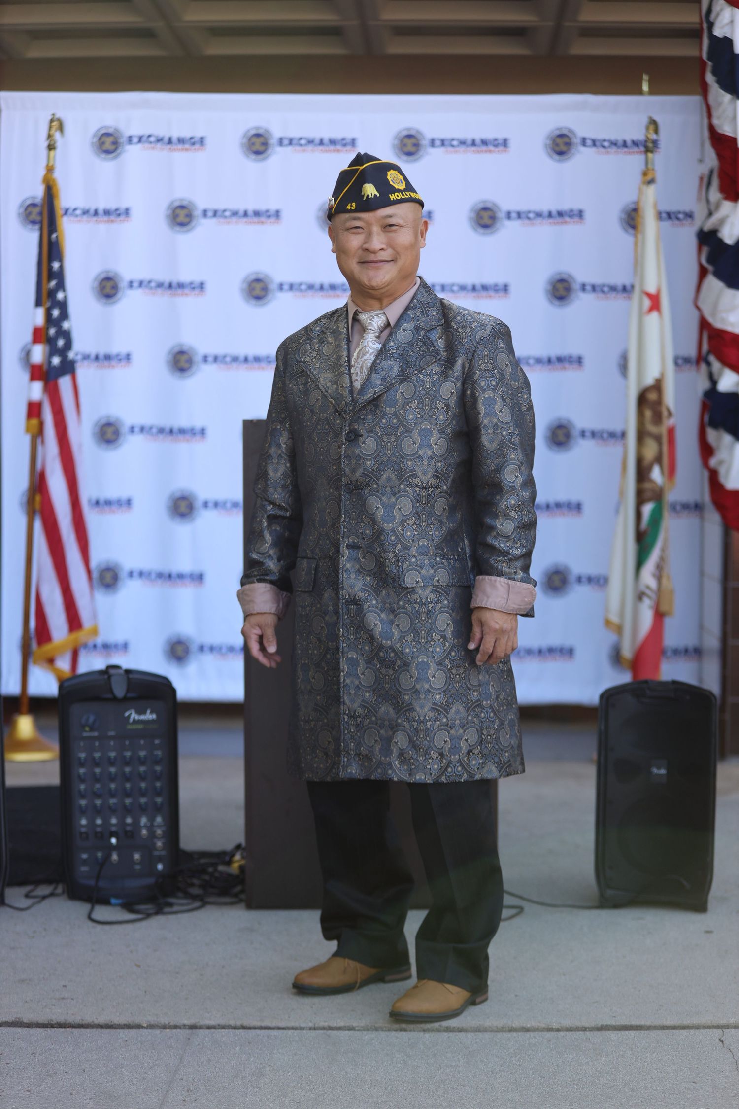 A person wearing a gray patterned jacket and dark pants stands in front of a branded backdrop with flags.