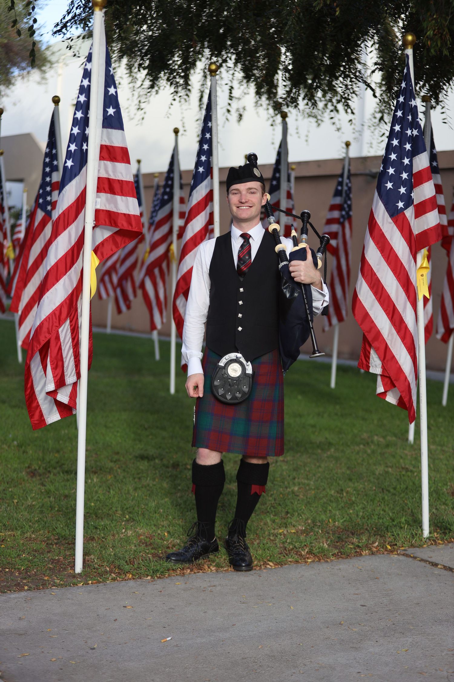 Person in formal Scottish attire poses with American flags displayed on lawn.