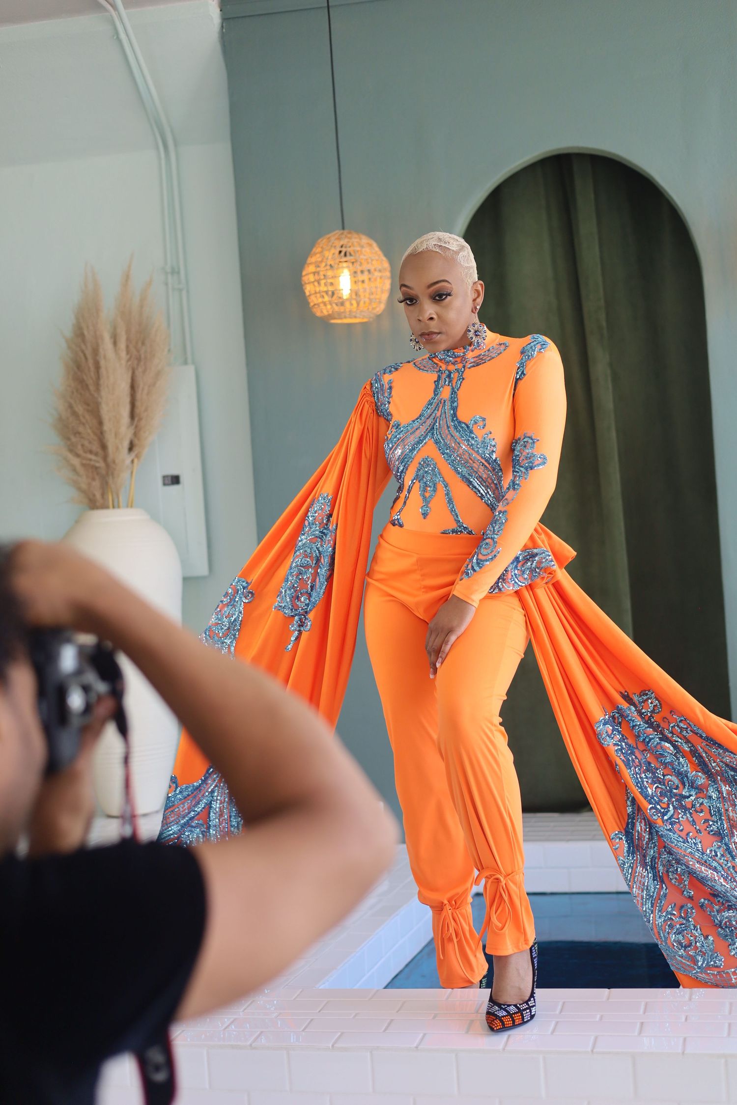 Model in bright orange dress with blue accents poses during a photoshoot against light blue wall.