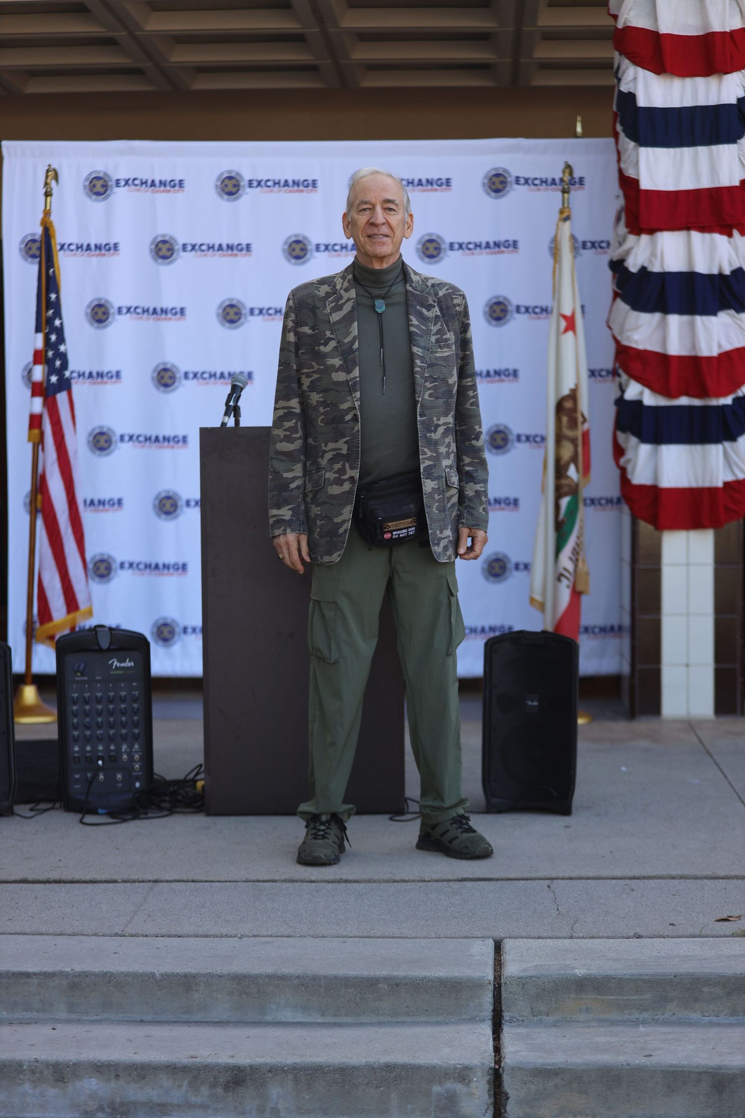 Military style outfit with camouflage jacket and olive pants on display stage with American flag.