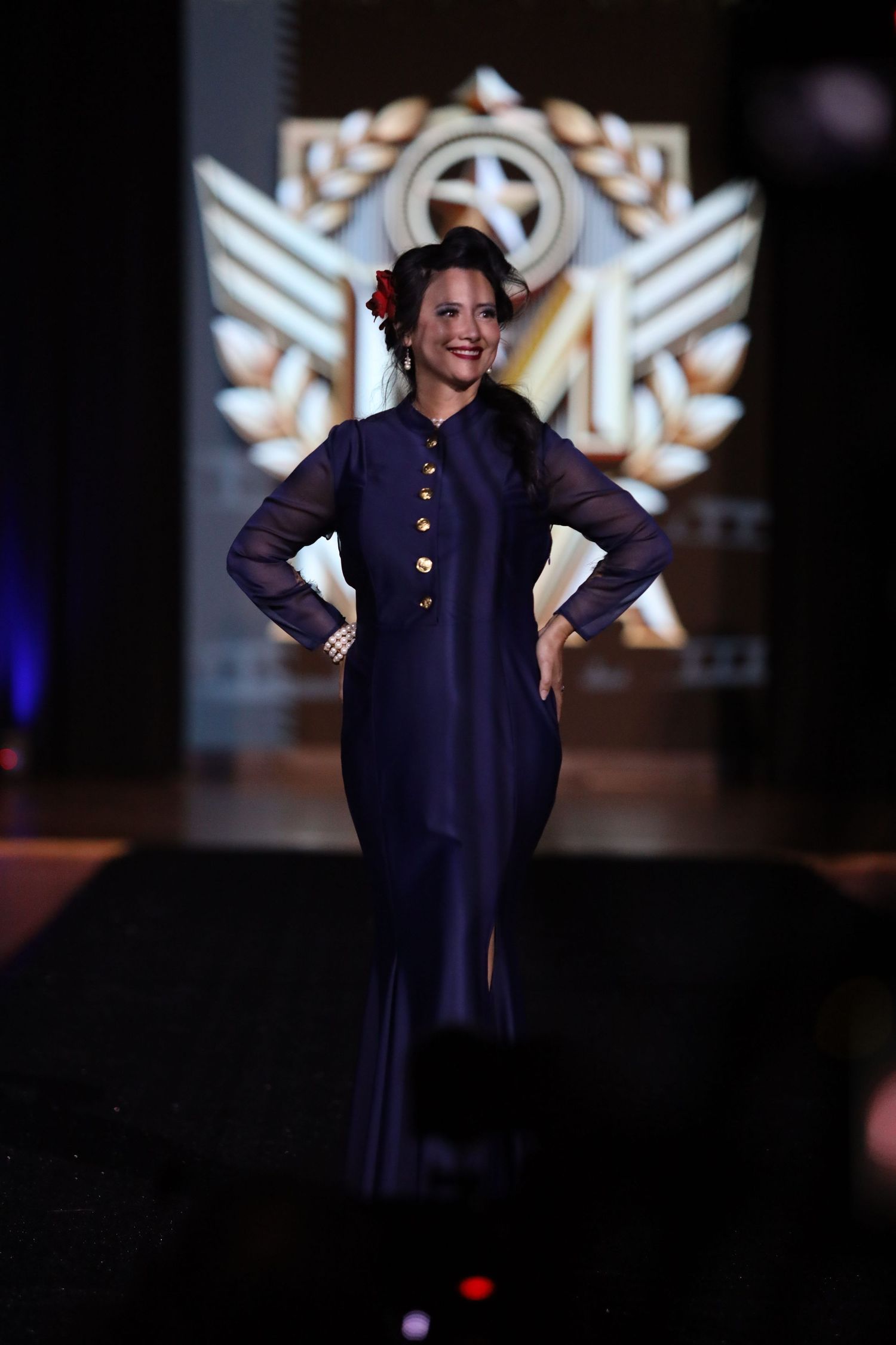 Fashion show model walks runway in navy military-inspired dress against dark background.