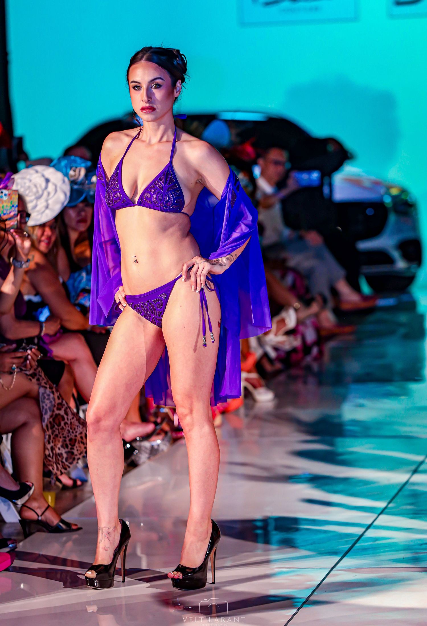 Model presents purple bikini with sheer cover-up on illuminated fashion runway.