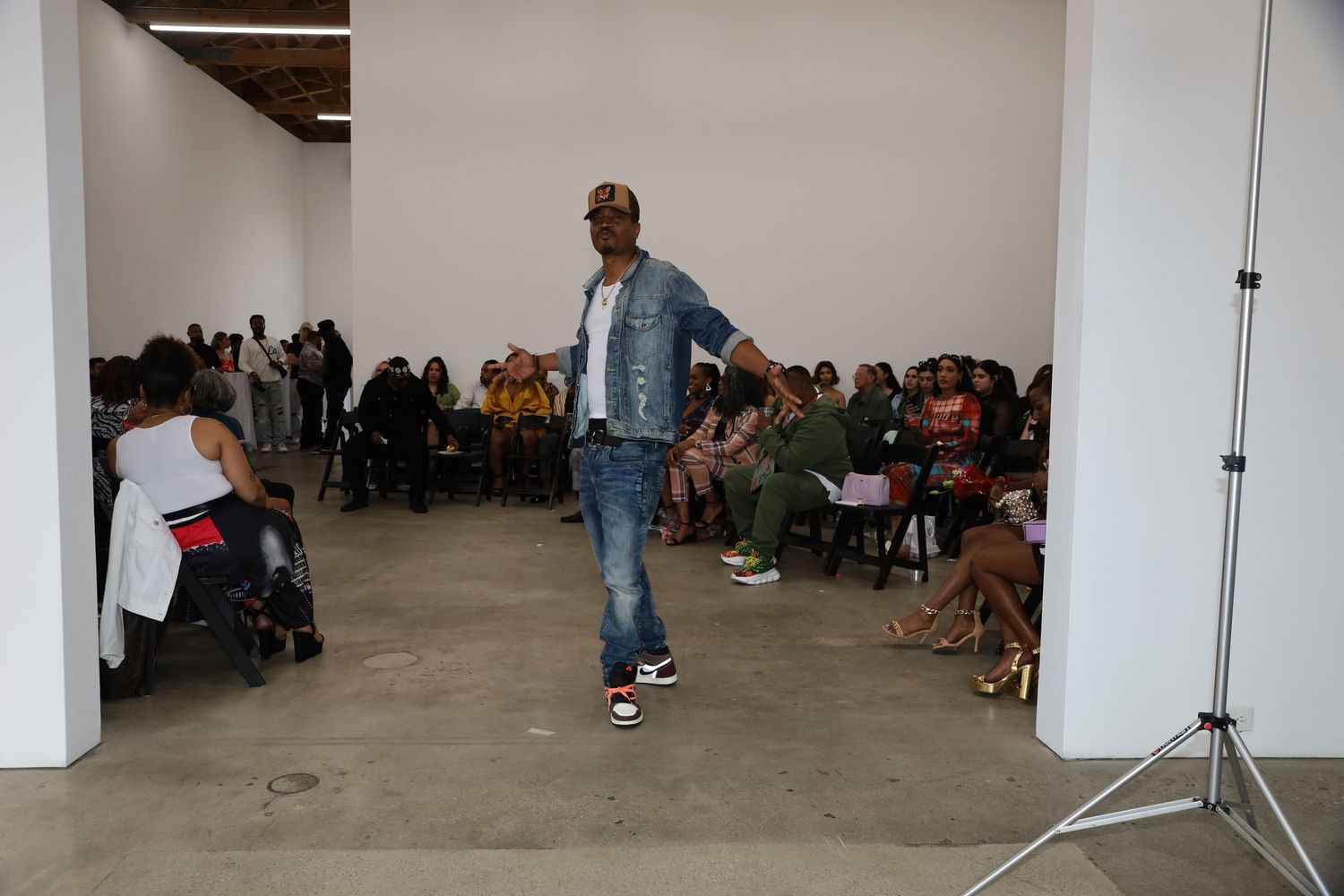 Fashion model showcasing streetwear on concrete runway with seated audience in background.