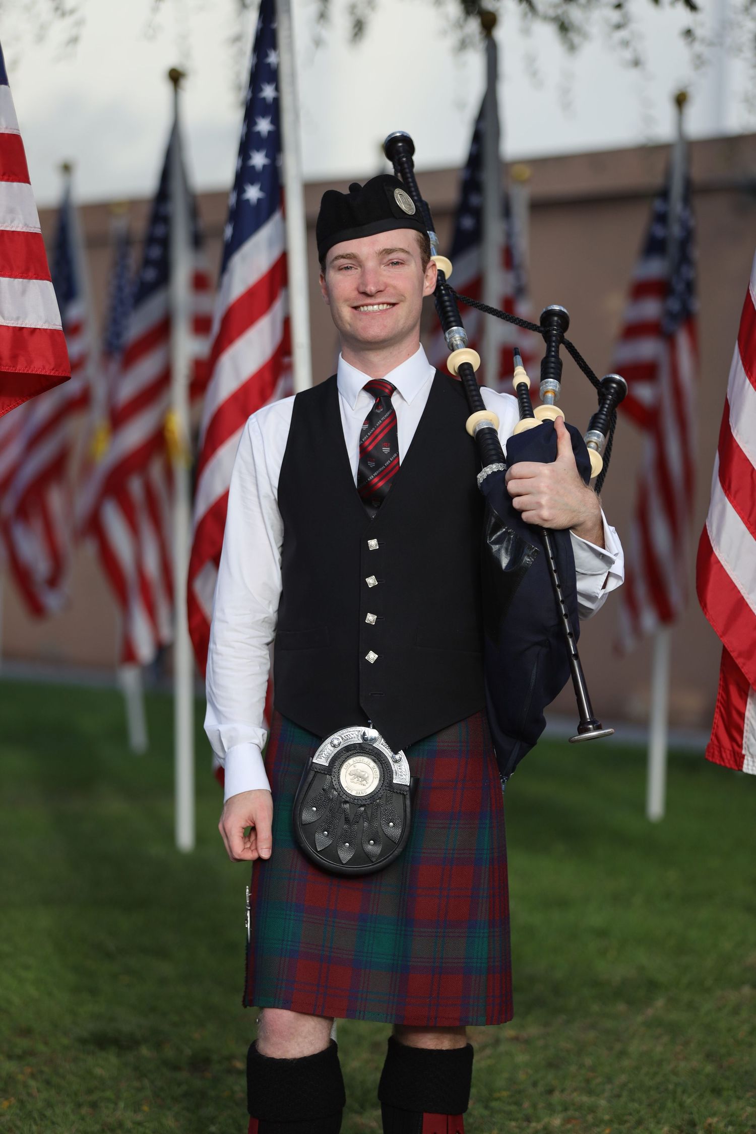Person in traditional Scottish kilt and formal attire poses with American flags in background.