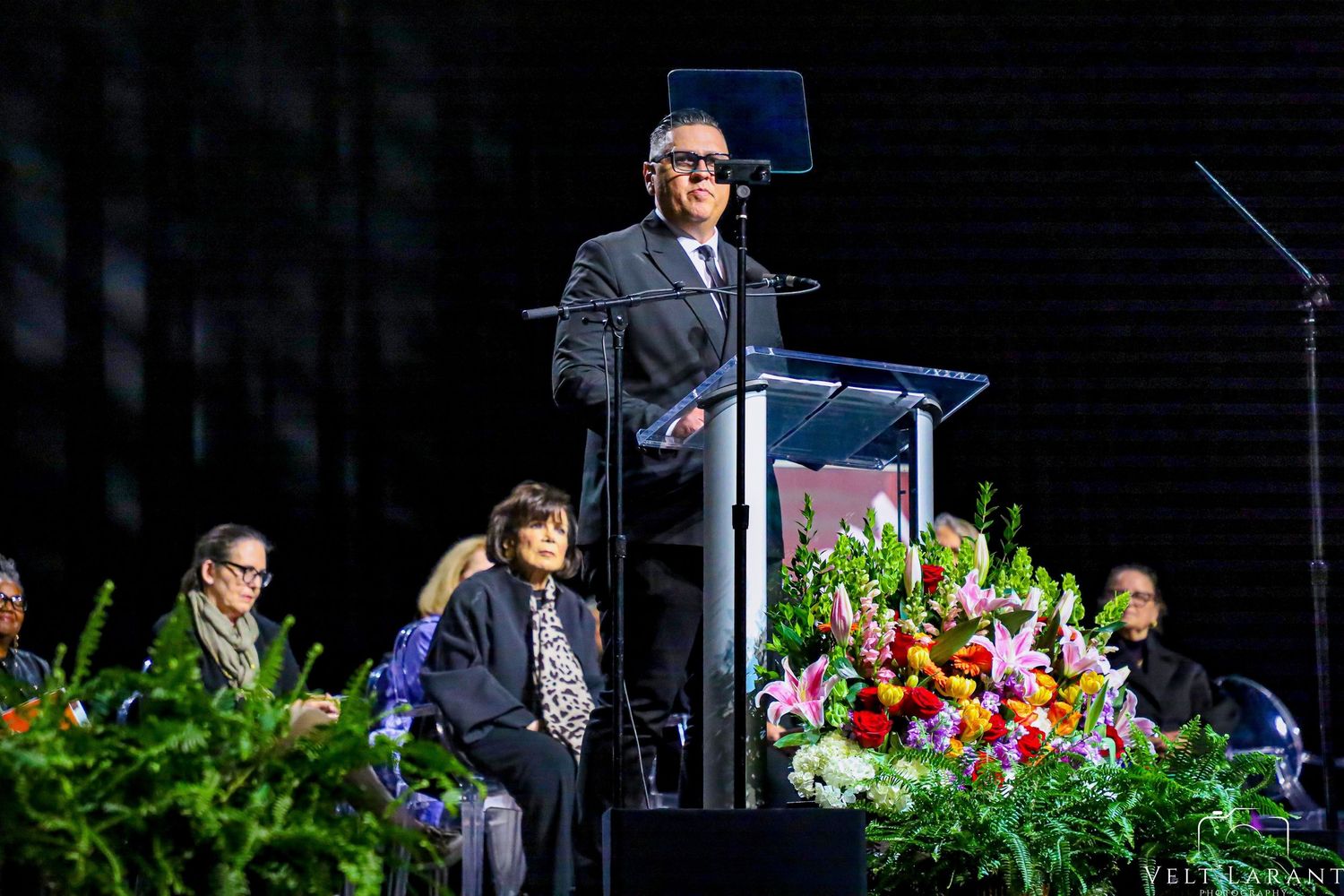 Speaker delivers address from illuminated podium with floral arrangements at graduation ceremony.