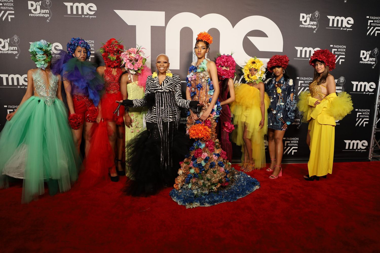 Red carpet event featuring multiple people wearing vibrant formal attire against a dark branded wall.