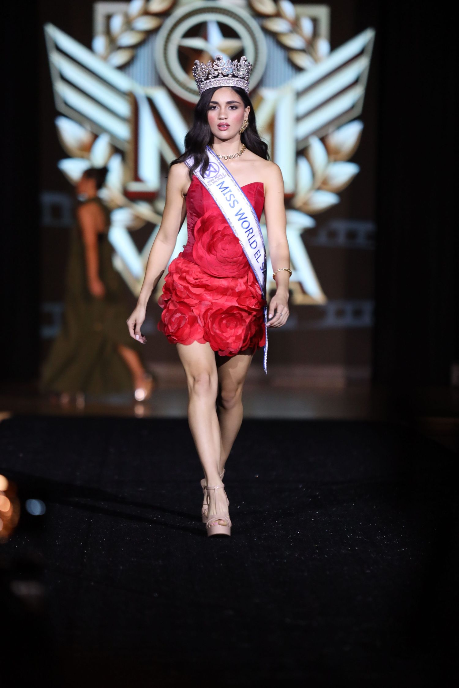Pageant contestant walks runway in red mini dress and crown against illuminated backdrop.