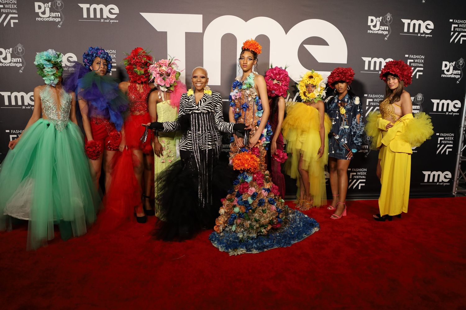 Fashion event attendees lined up in colorful evening wear against a branded step-and-repeat wall.