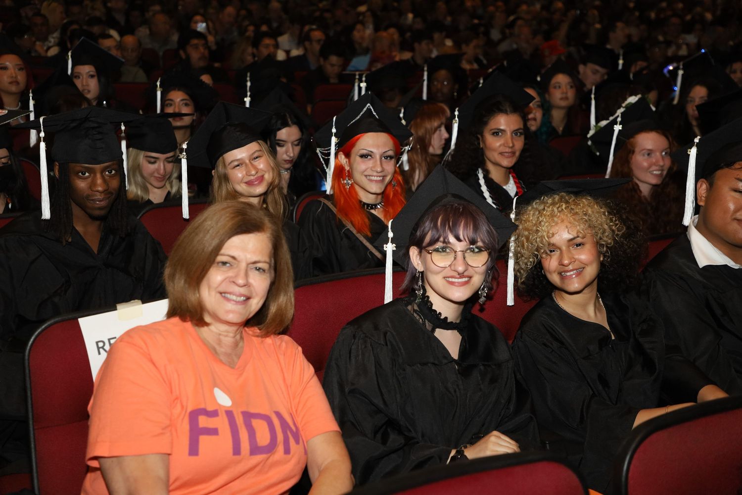 Graduation ceremony audience members seated in theater with person wearing orange shirt visible.