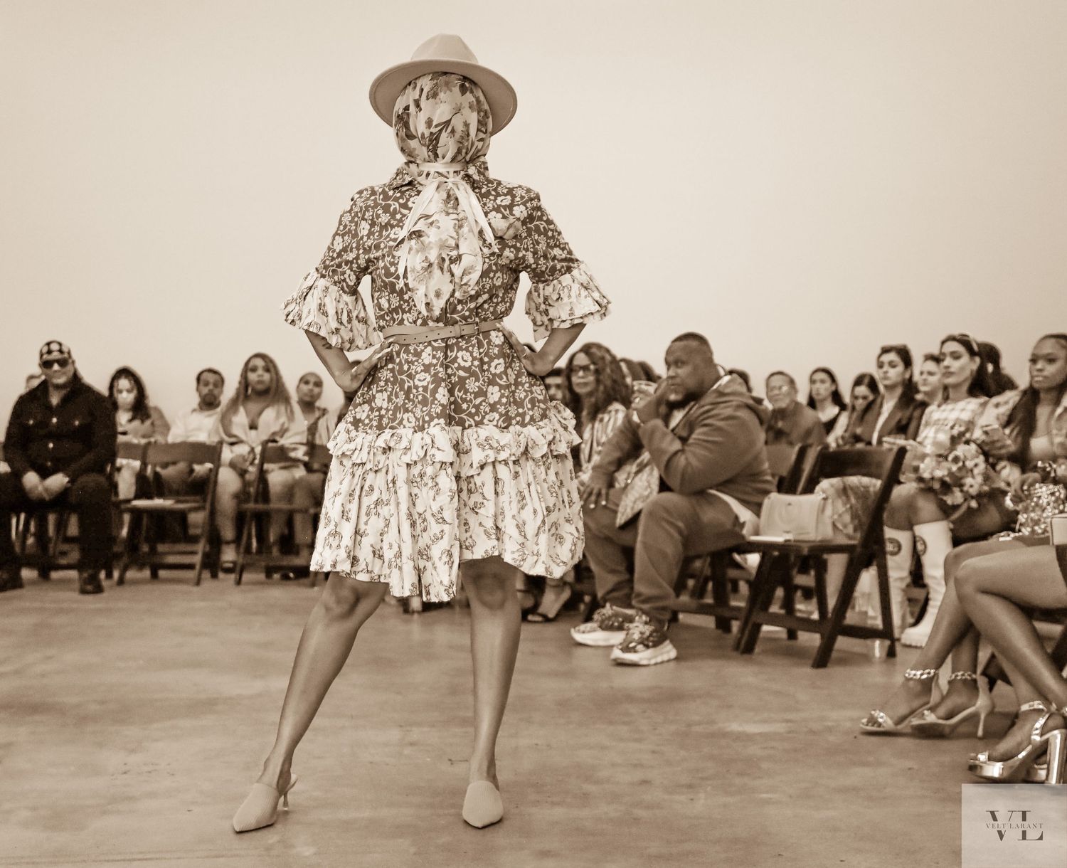 Vintage sepia photo of a performer in a patterned dress and hat dancing in front of a seated audience.