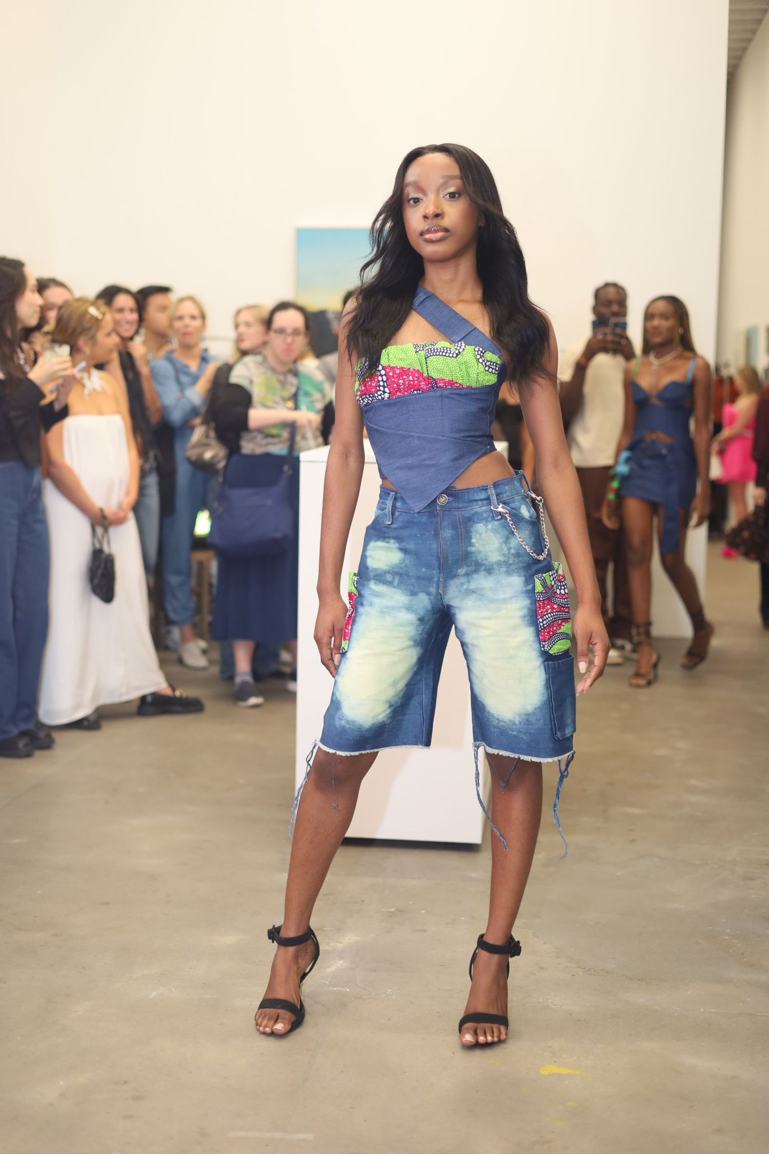 Model poses in denim capris and colorful crop top on runway with crowd in background.