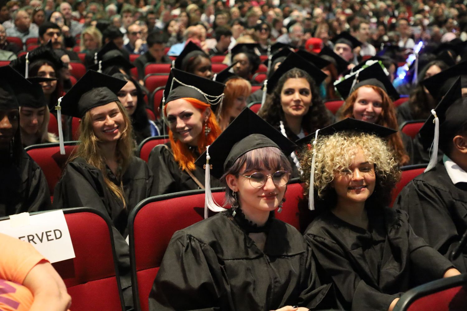Wide shot of packed auditorium during graduation ceremony with graduates in black robes.