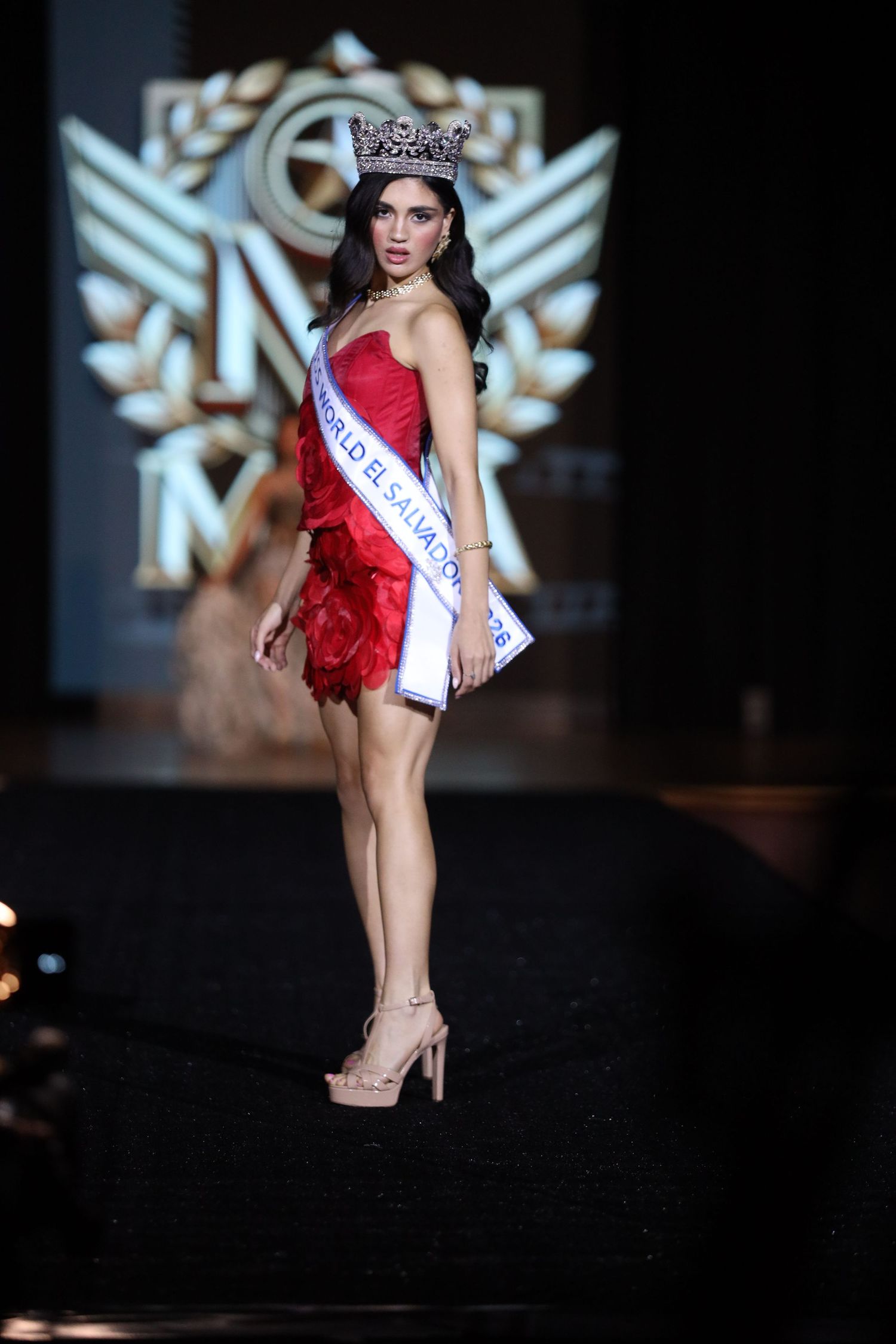 A pageant contestant in a red mini dress and crown poses on stage with a sash during a fashion show competition.